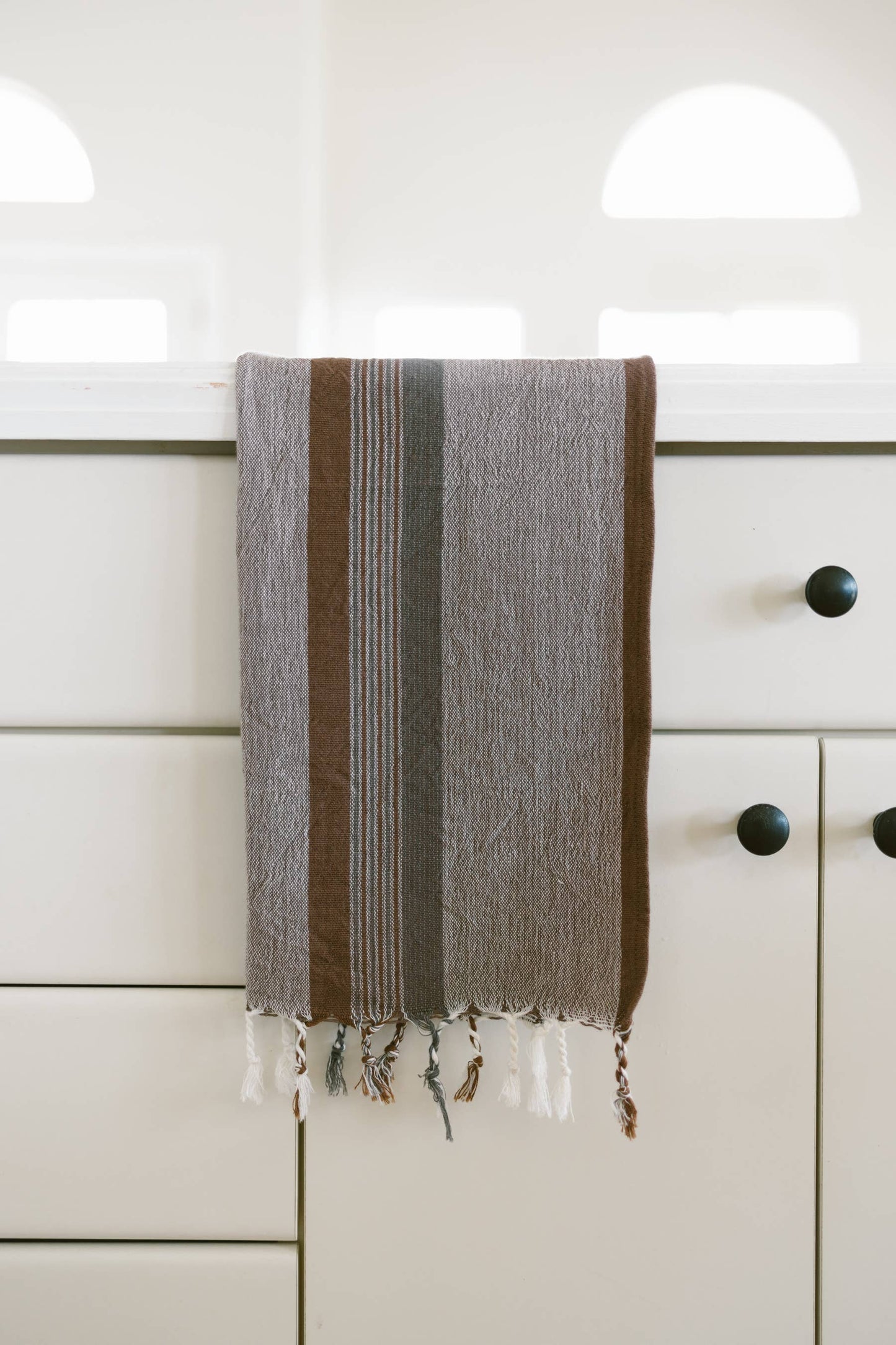 Grey, white, and dark brown cotton kitchen towel with fringe hangs on the door of a white kitchen cabinet with black knobs