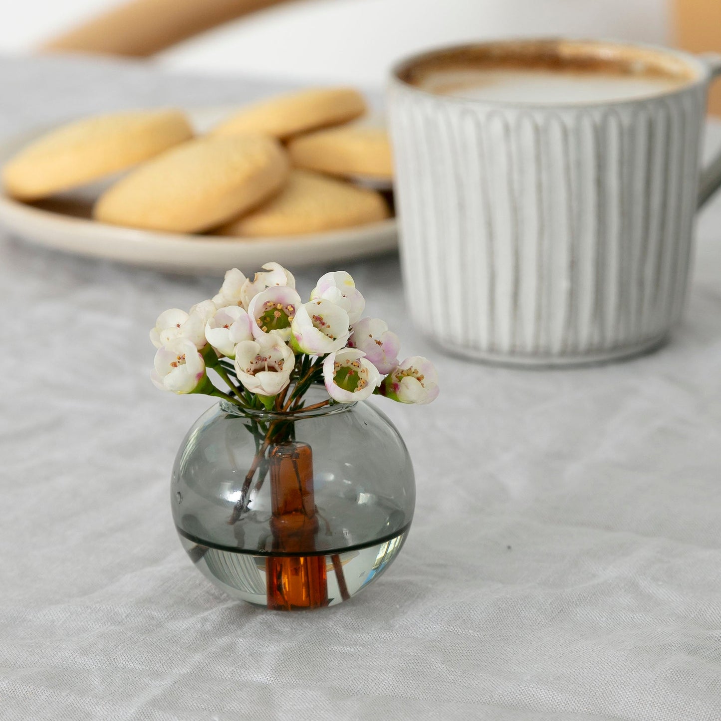 Spherical smoky gray small vase with an orange glass tube in its interior is filled with small white flowers and sits on a table alongside a plate of shortbread cookies and a cup of coffee
