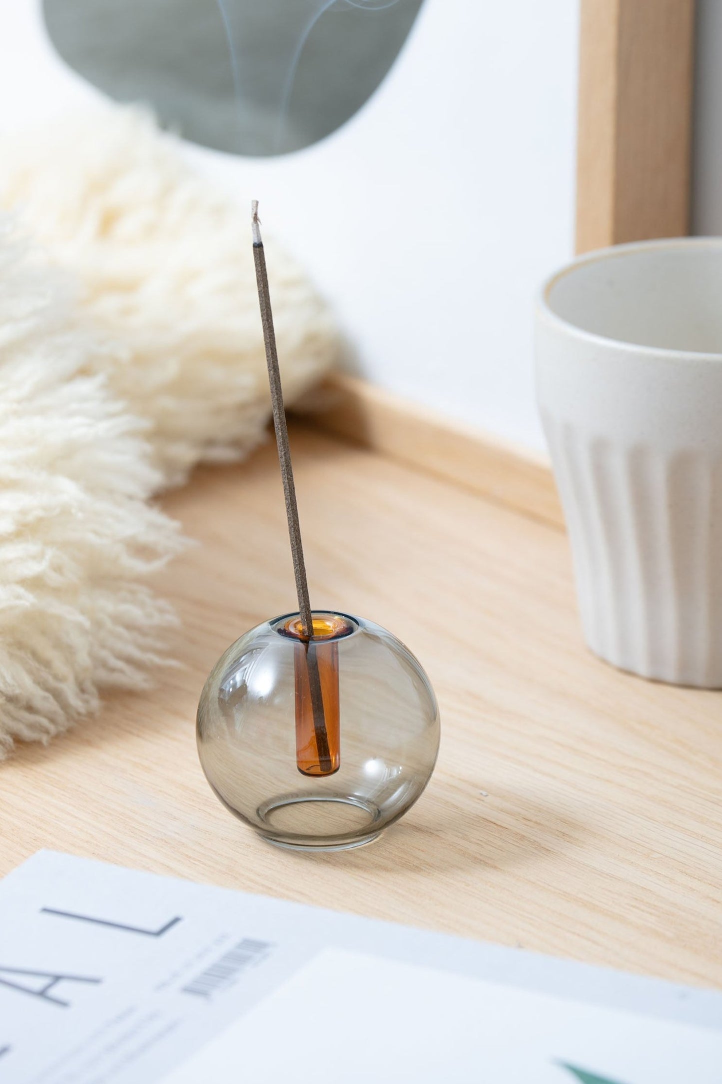 Spherical smoky gray small vase with an orange glass tube sits on a light wood desk holding an incense stick with a lambskin pillow and magazine beside it