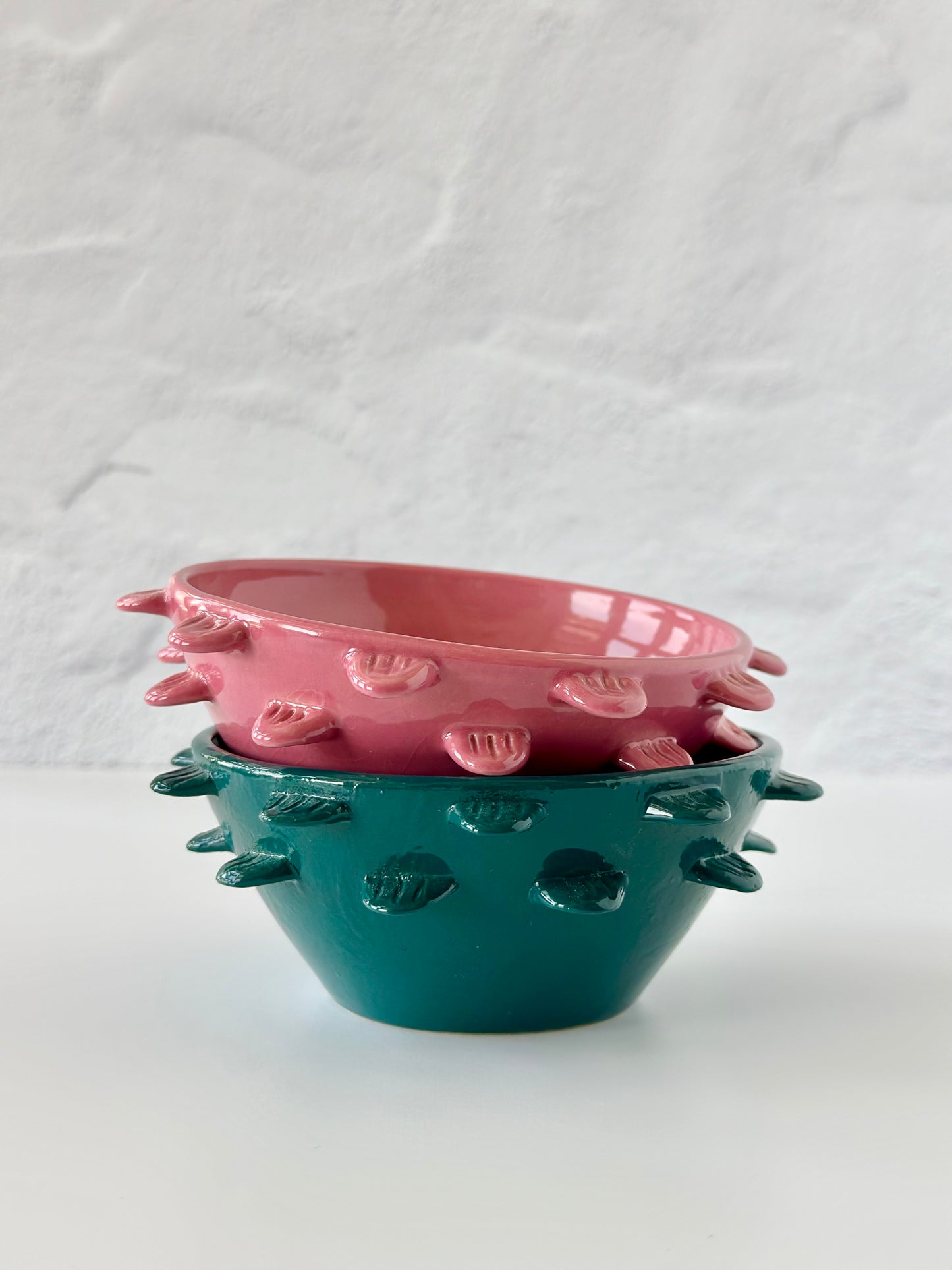 Two ceramic bowls, one pink and one green, with feather-like details on their exterior, stacked together against a textured background.