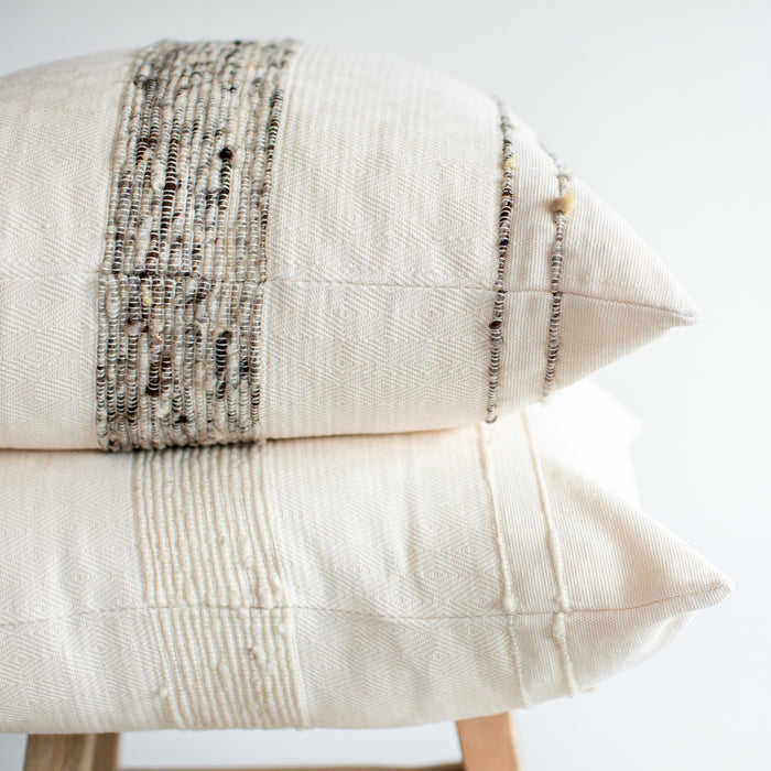 Two pillows laid on on top of one another, sitting on a wooden stool - both pillows have ivory wool and cotton covers with striping detail, one in ivory, the other in dark grey