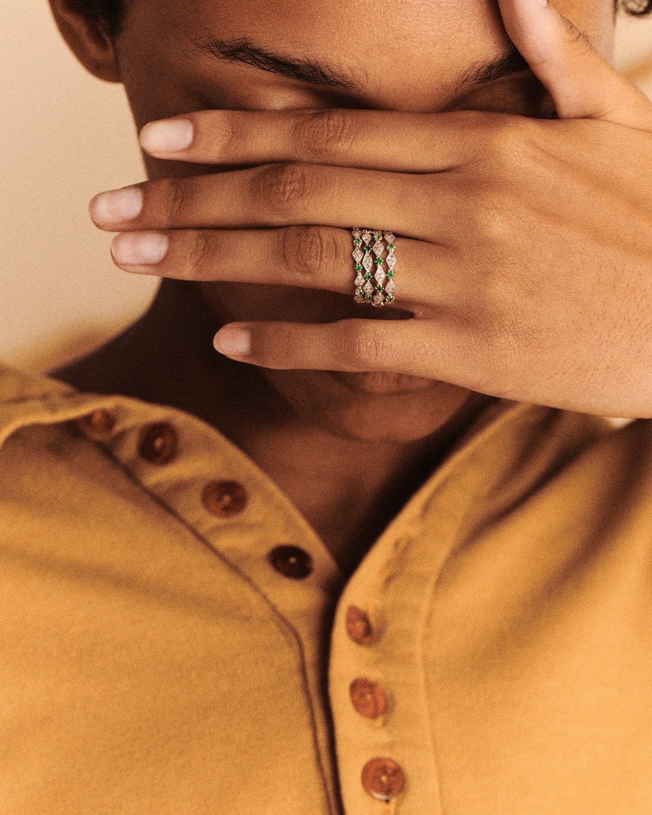 A woman with dark eyebrows in a golden yellow buttoned shirt partially covers her face with her left hand, showing a stack of four gold, diamond, and emerald thin rings with diamond-shaped motif on her ring finger