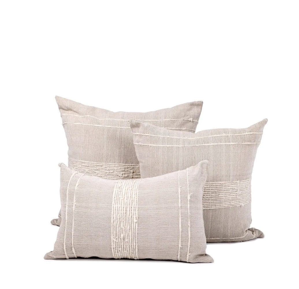 Trio of multi-sized square and rectangular pillows with sand-colored wool and cotton covers featuring woven ivory striping details sits against white background