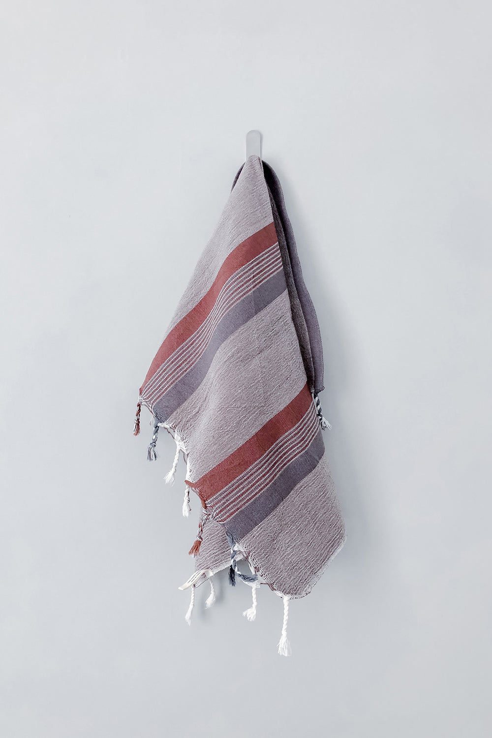 Grey, white, and dark brown cotton kitchen towel with fringe hangs from a hook against a white background
