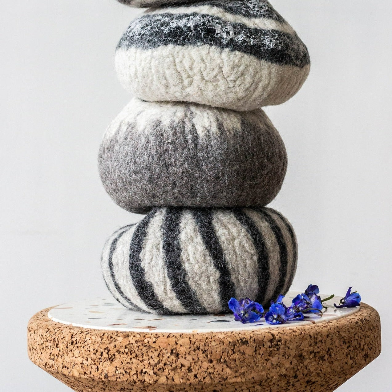 A trio of ivory and grey wool decorative bowls sits in a stack on a cork and stone pedastal table with some purple-blue small flowers strewn to the side