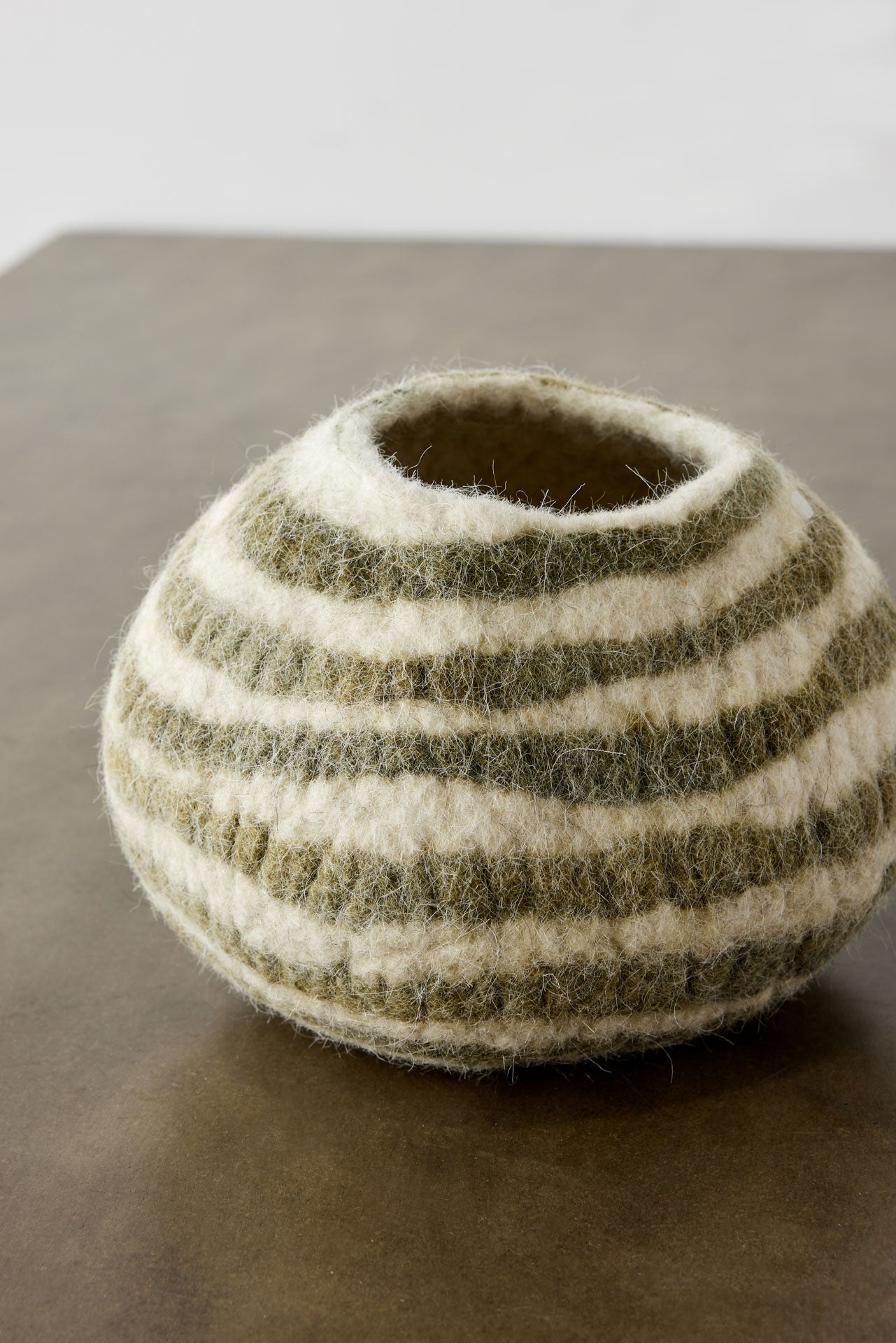 A close-up, partial view of an ivory wool decorative bowl with olive green stripes sits on a wooden surface with a white background