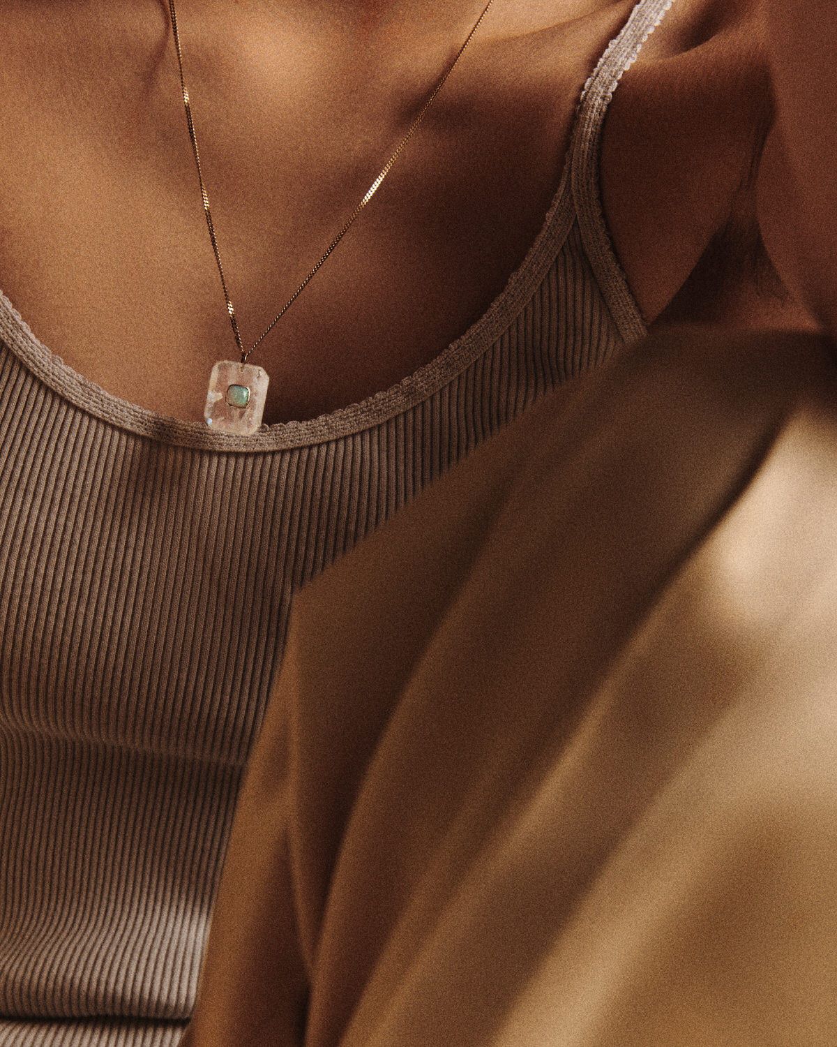 The partial chest of a woman wearing a beige ribbed tanktop and with her knee drawn up against her body wearing gold satin pants and a gold chain necklace with a rectangular moonstone pendant that features a round opal in the center set in a ring of yellow gold
