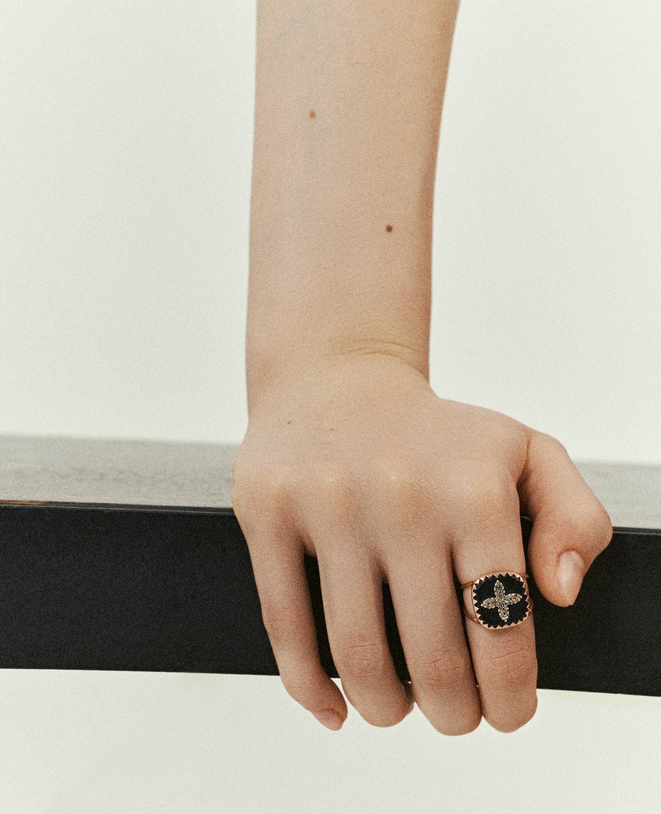 Woman's wrist and hand holding a black railing with a double-banded rose gold ring with square black resin medallion featuring a four-petal shape diamond inlay on her pointer finger