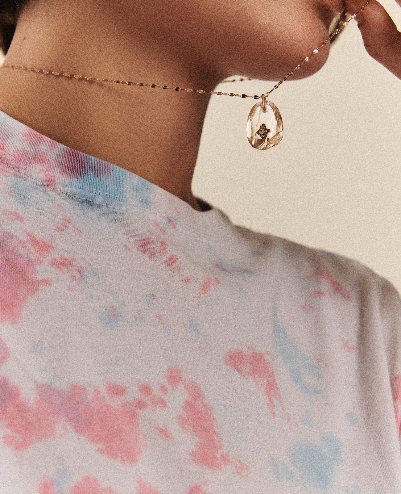 The view of the chest, shoulders, neck and chin of a woman in a pink, white and blue tie-dyed tshirt wearing a twisted gold chain necklace with a subtly tear-drop shaped rutile quartz pendant with a diamond and gold inlay featured in the center that she is holding up to her face