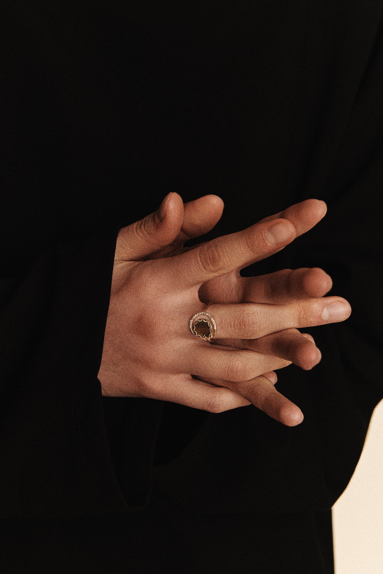 The hands of a woman in a black top showing a gold ring with a round shape tucked into a half-ring of diamonds set against an off-white background worn on her middle finger