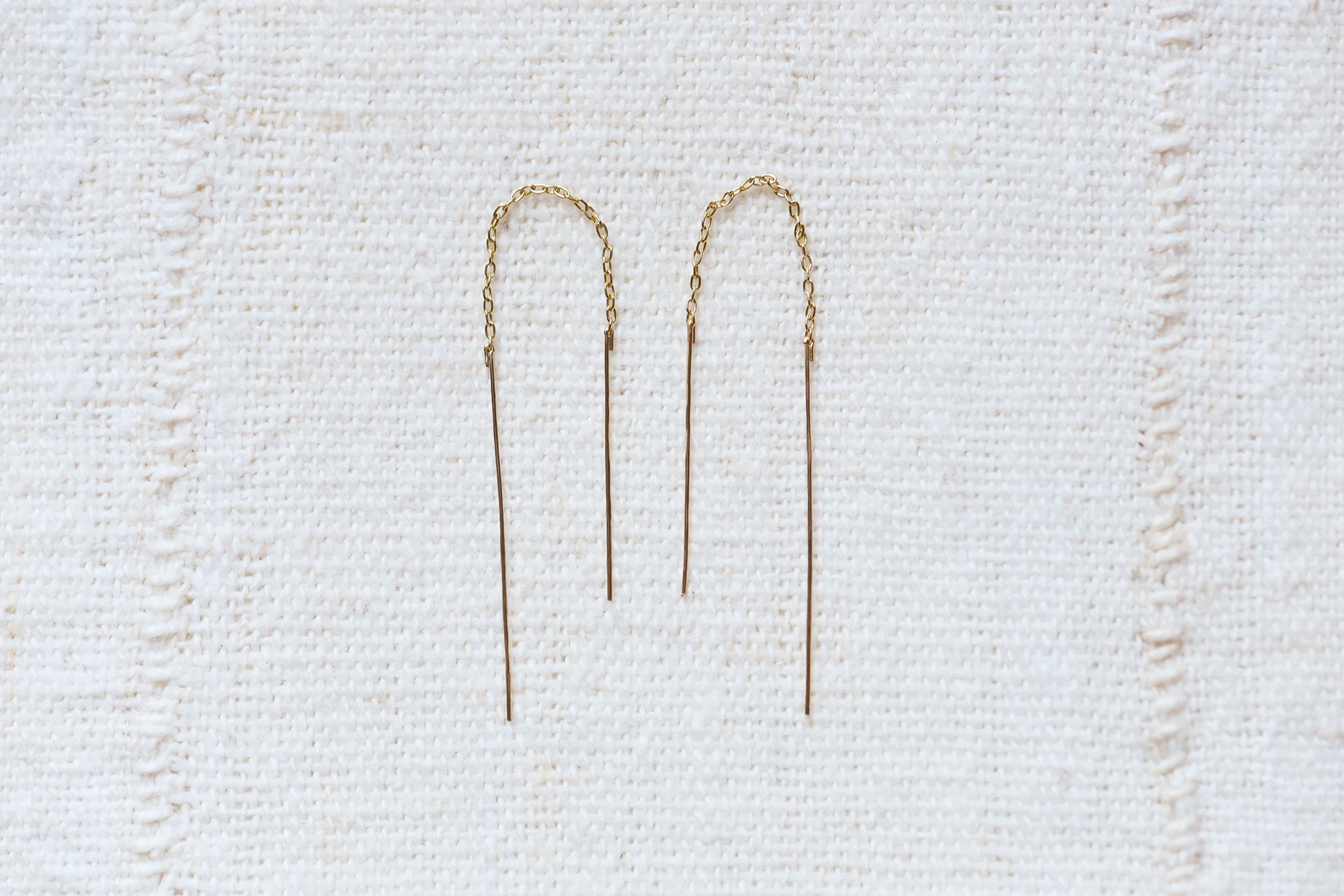 Two gold threader earrings on a textured white cloth background