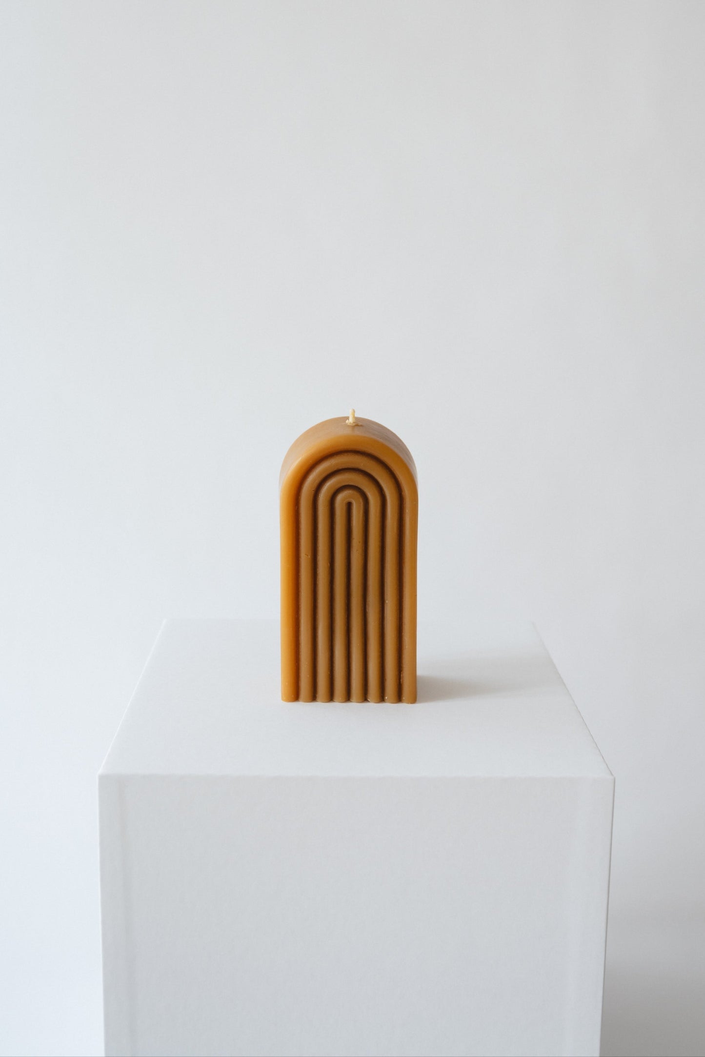 A hand-poured beeswax candle with a golden rainbow design, displayed on a white pedestal against a neutral background.