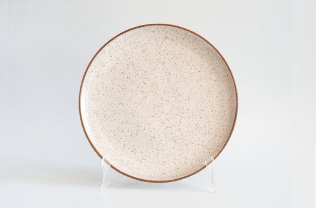 A round ceramic serving platter with a speckled beige and white design, displayed on a white stand against a white background.