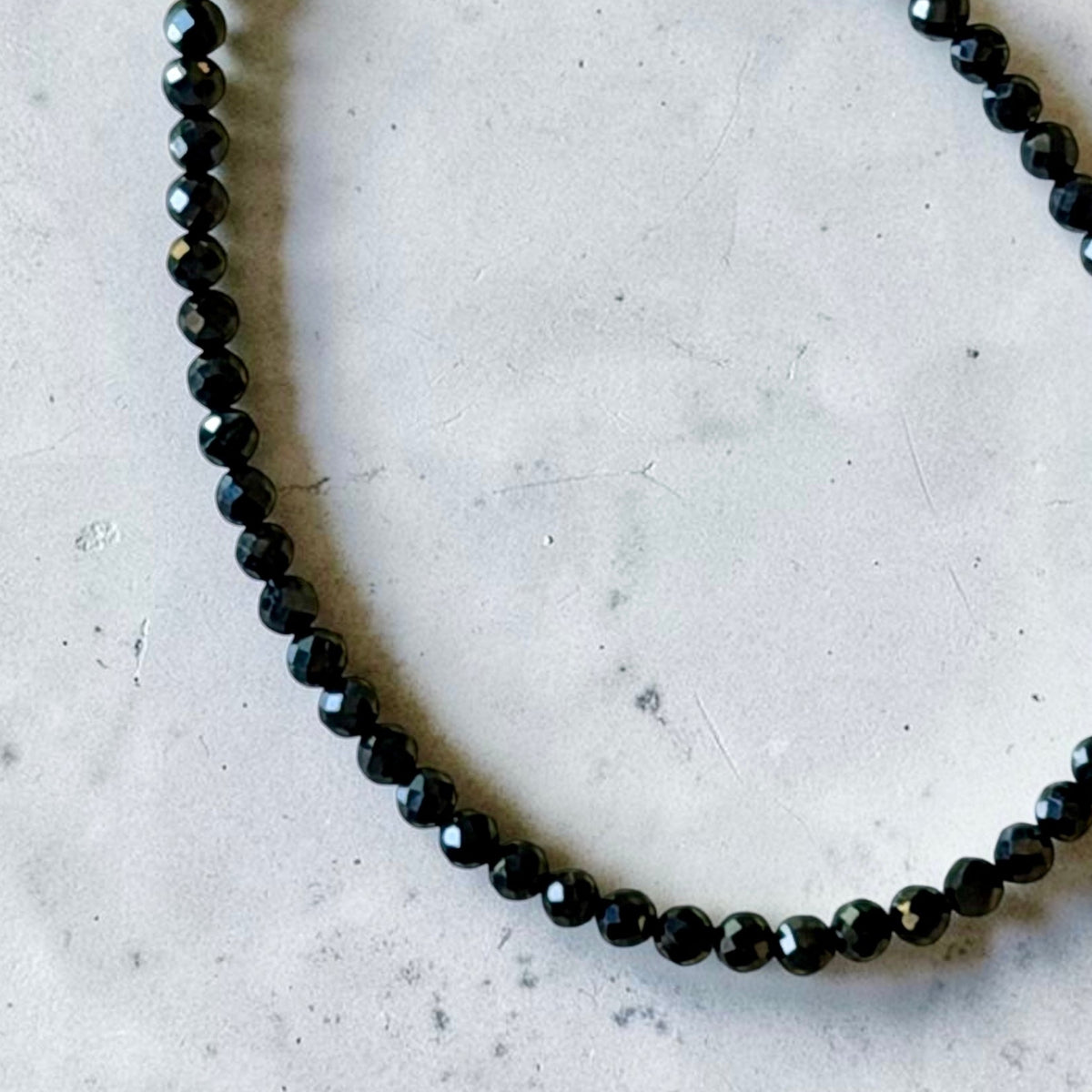 A beaded bracelet made of small faceted black beads with a gold-tone lobster clasp, photographed on a light gray stone-like surface.