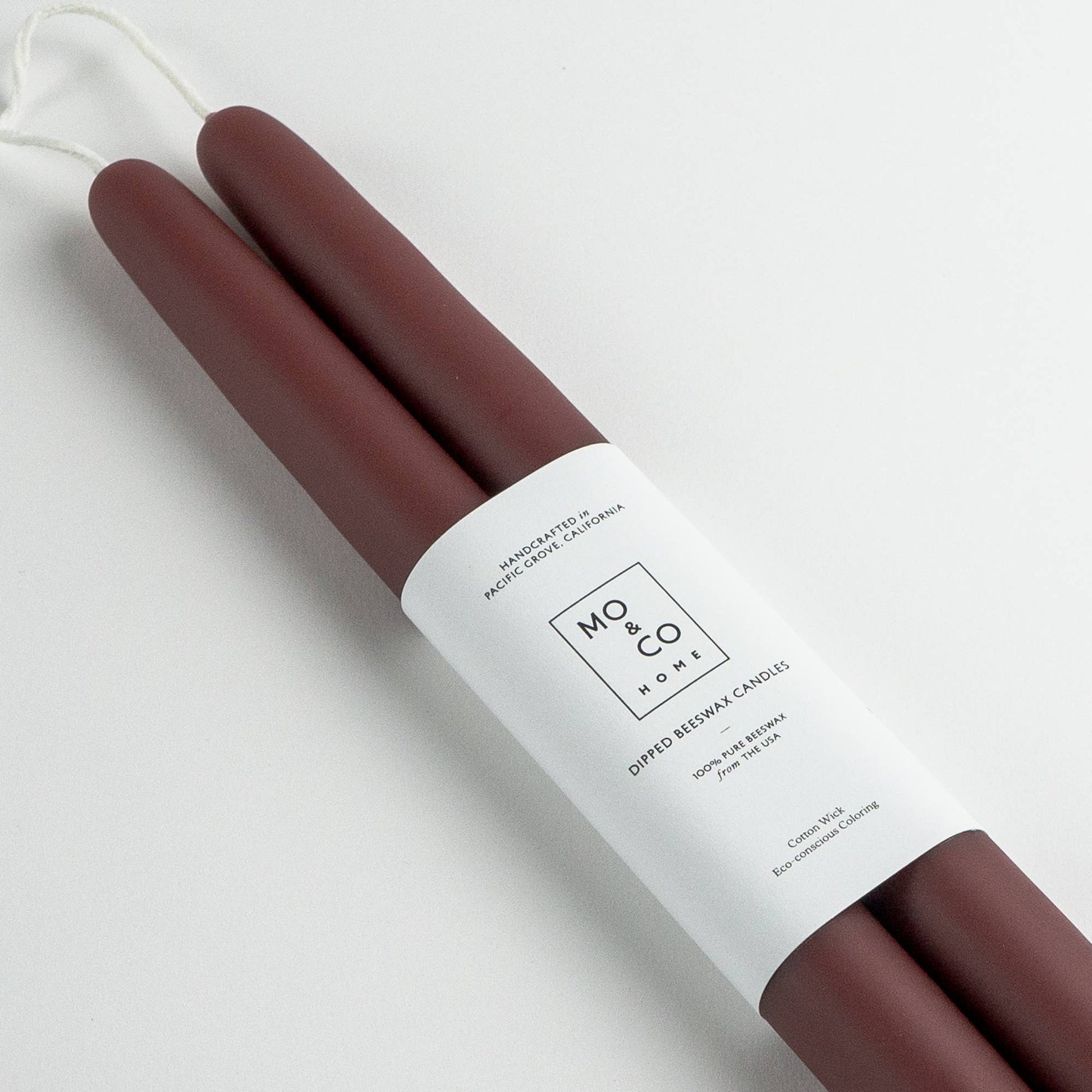 Pair of burgundy beeswax taper candles with a white label on a light gray background