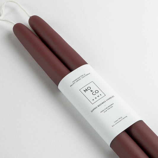 Pair of burgundy beeswax taper candles with a white label on a light gray background