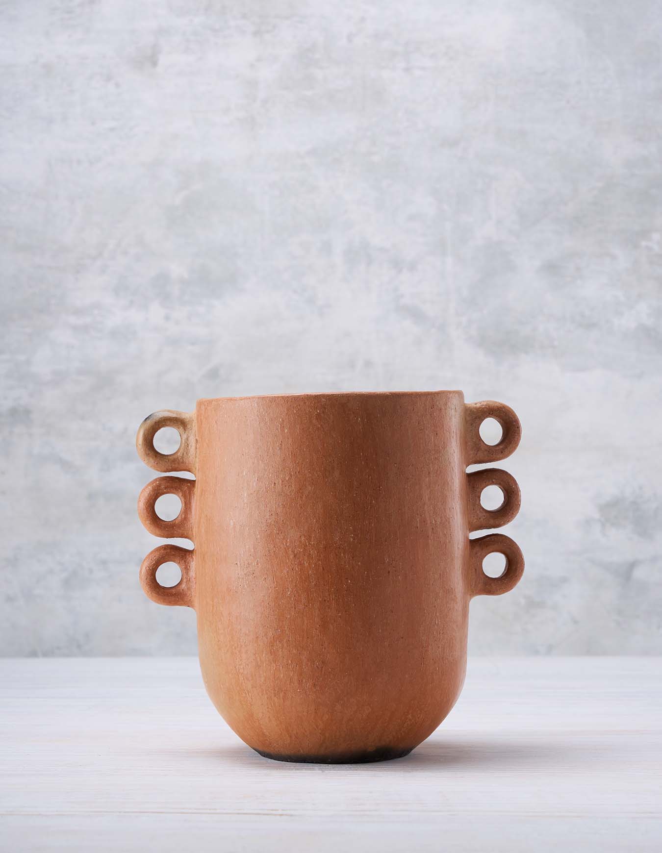 Clay vessel in terracotta color with ring accents on both sides set on a white surface against a light grey background