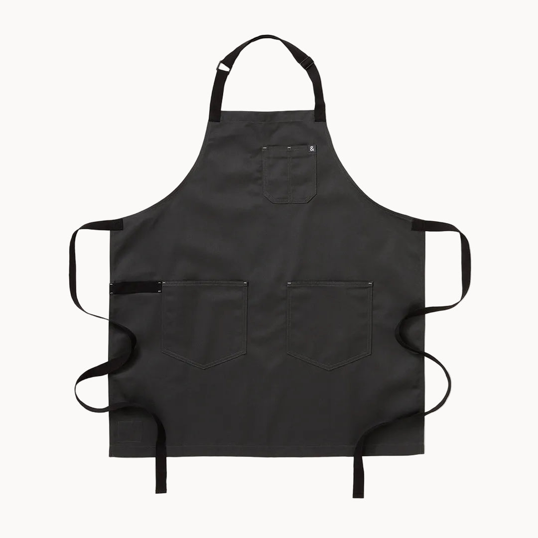 A black bib apron with four front pockets and adjustable neck and waist straps, laid flat against a light gray background.