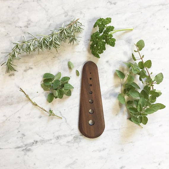 Walnut wood herb stripper sitting on a white marble surface surrounded by green, leafy herbs 