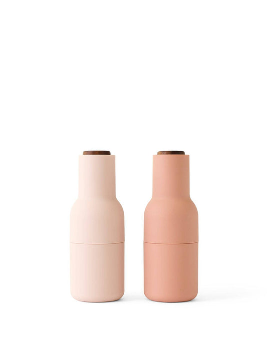 Two cylindrical spice bottle grinders with a bottle-shaped design, one in a lighter blush color and the other in a darker nude tone, both with walnut wood lids and a silicone coating.