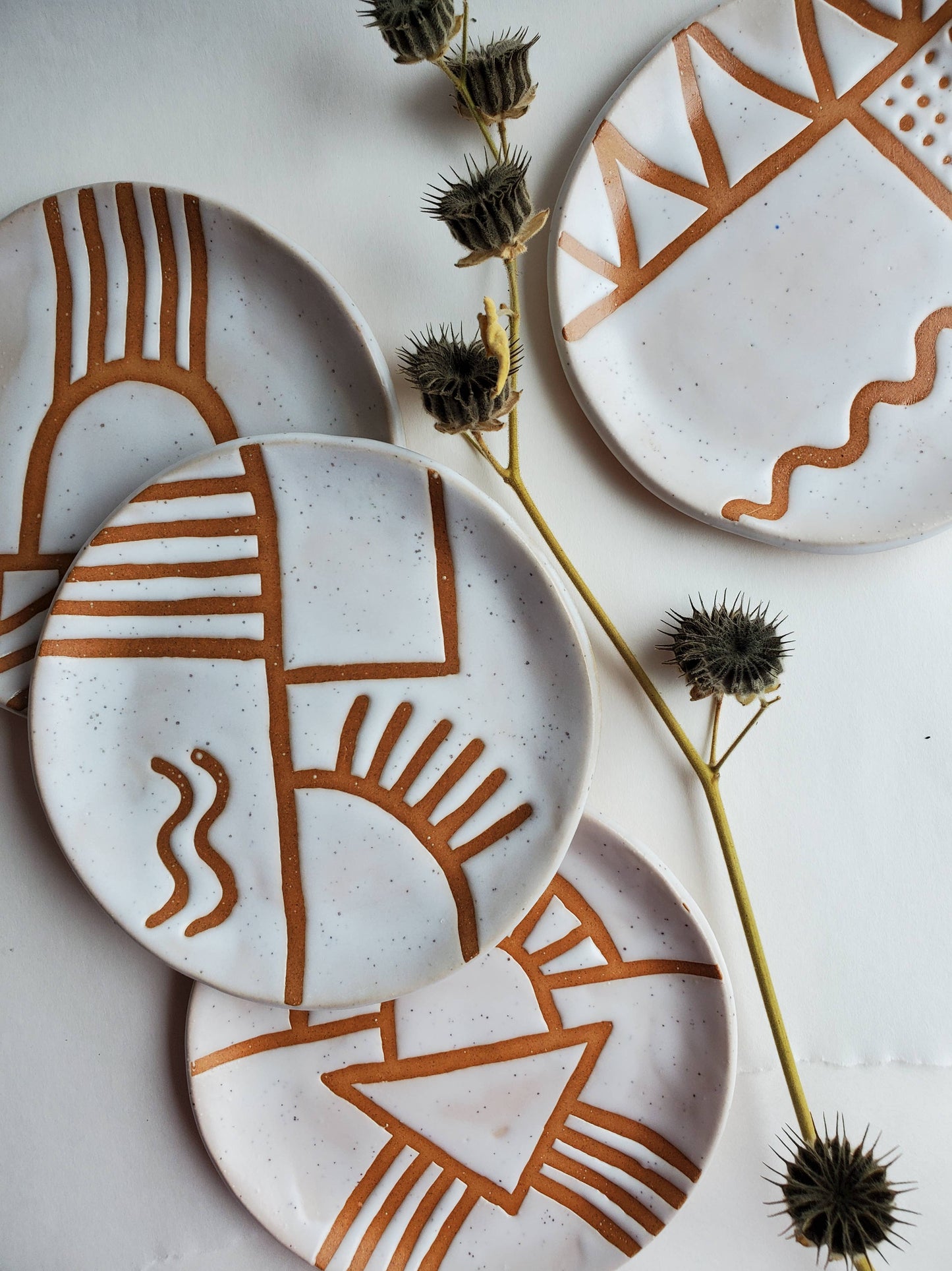 A set of white ceramic plates with geometric brown doodles, displayed on a table with natural lighting and a plant in the background.