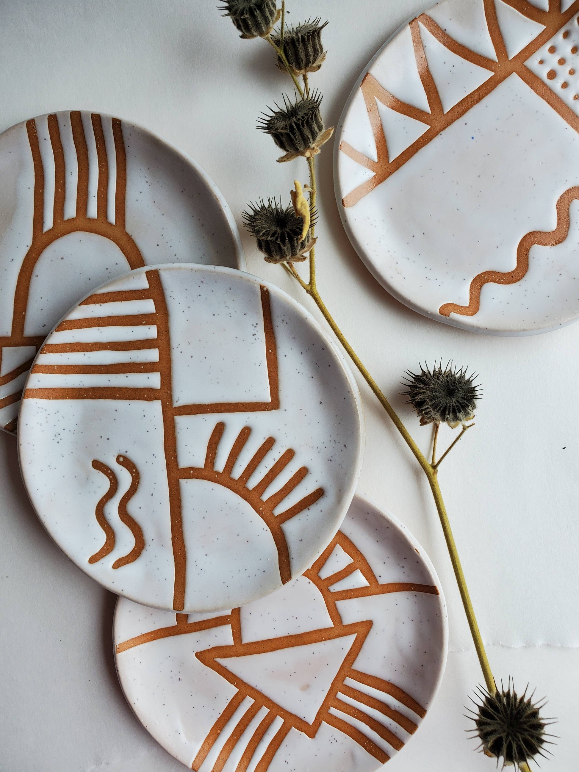 A set of white ceramic plates with geometric brown doodles, displayed on a table with natural lighting and a plant in the background.