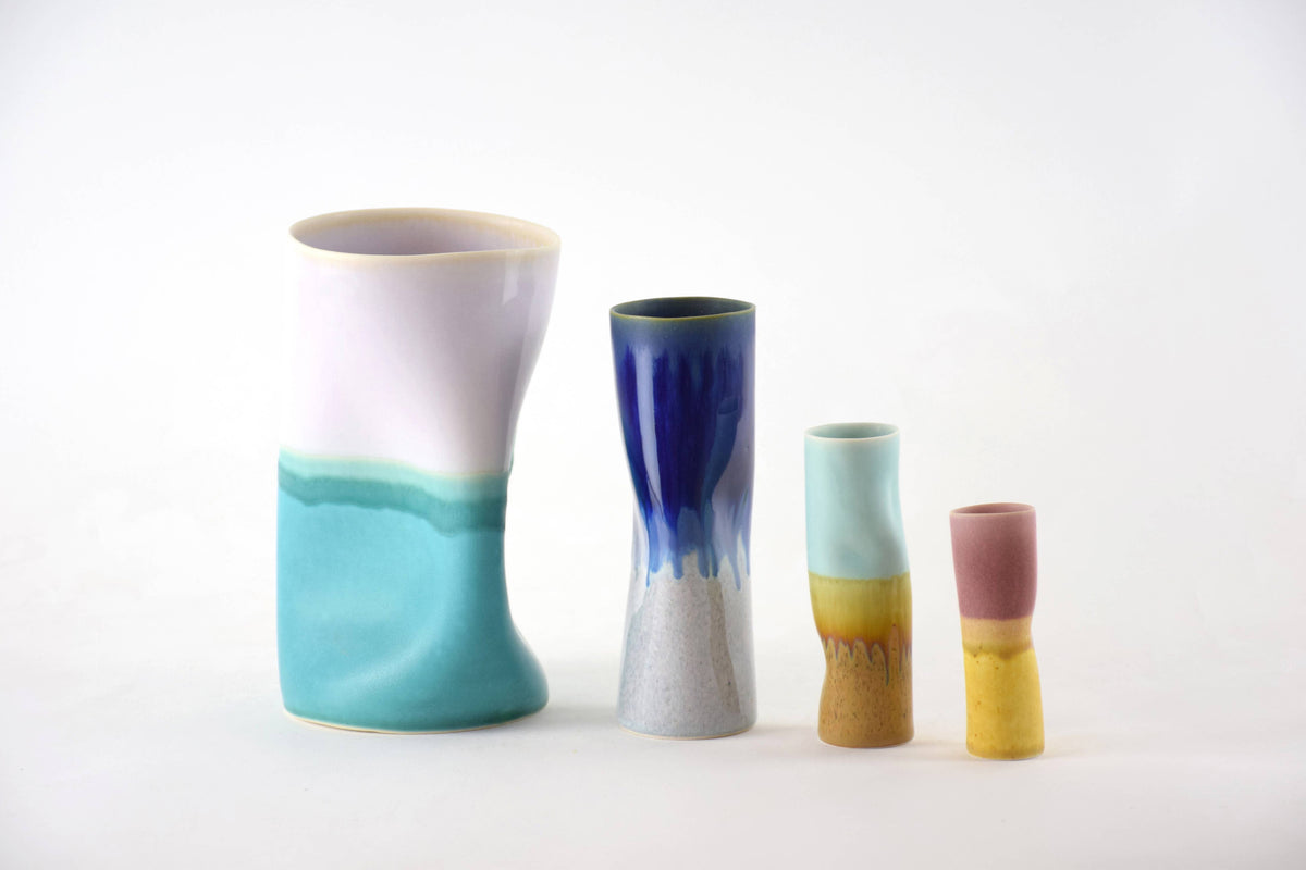 Four handmade ceramic vases of varying heights and shapes, featuring colorful glaze designs including pink and yellow, light blue and yellow, dark blue and white, and white and turquoise, arranged on a seamless white background.