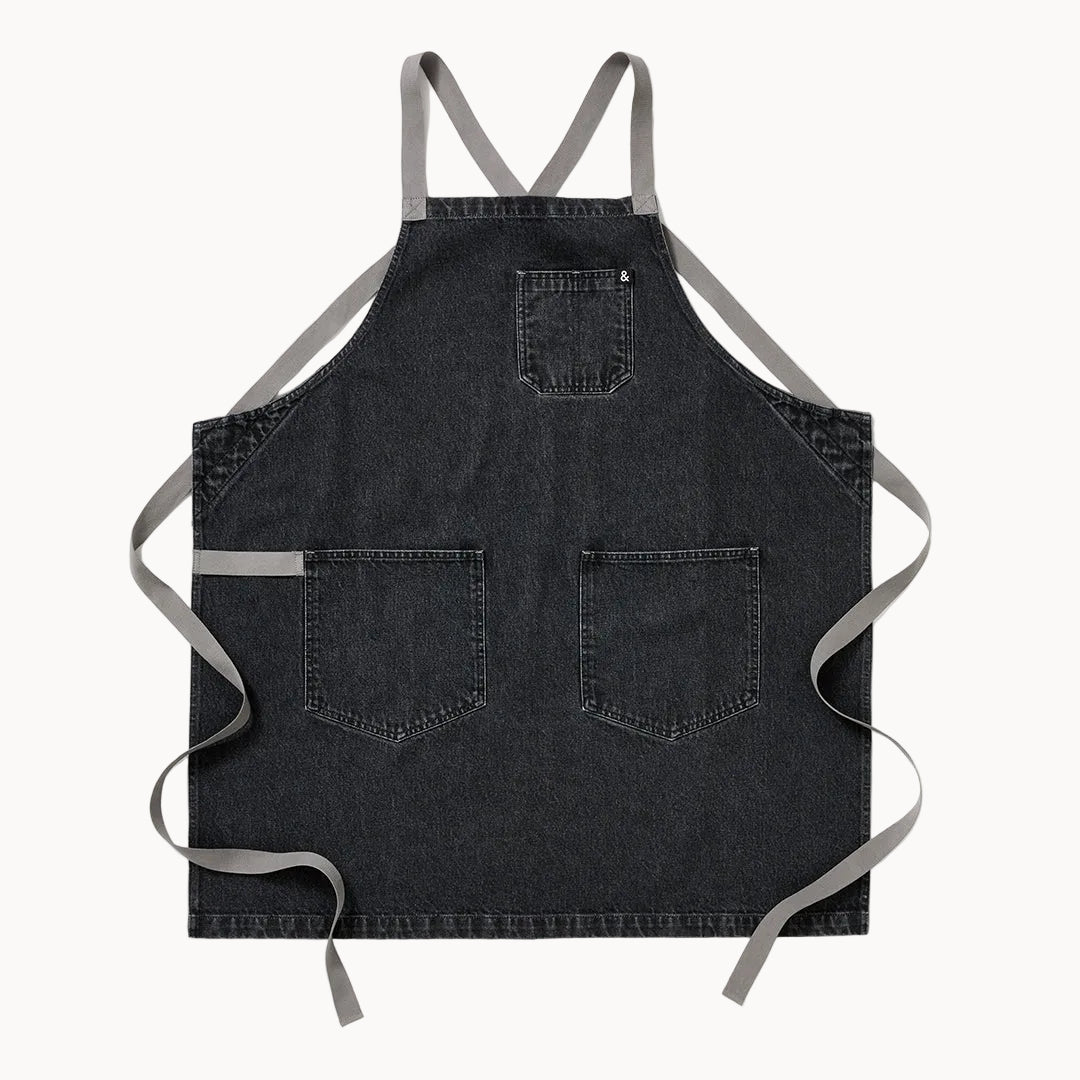 A dark gray denim apron with light gray straps, featuring two front pockets and a top bib pocket, laid flat against a white background.