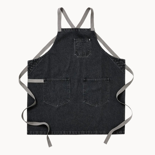 A dark gray denim apron with light gray straps, featuring two front pockets and a top bib pocket, laid flat against a white background.