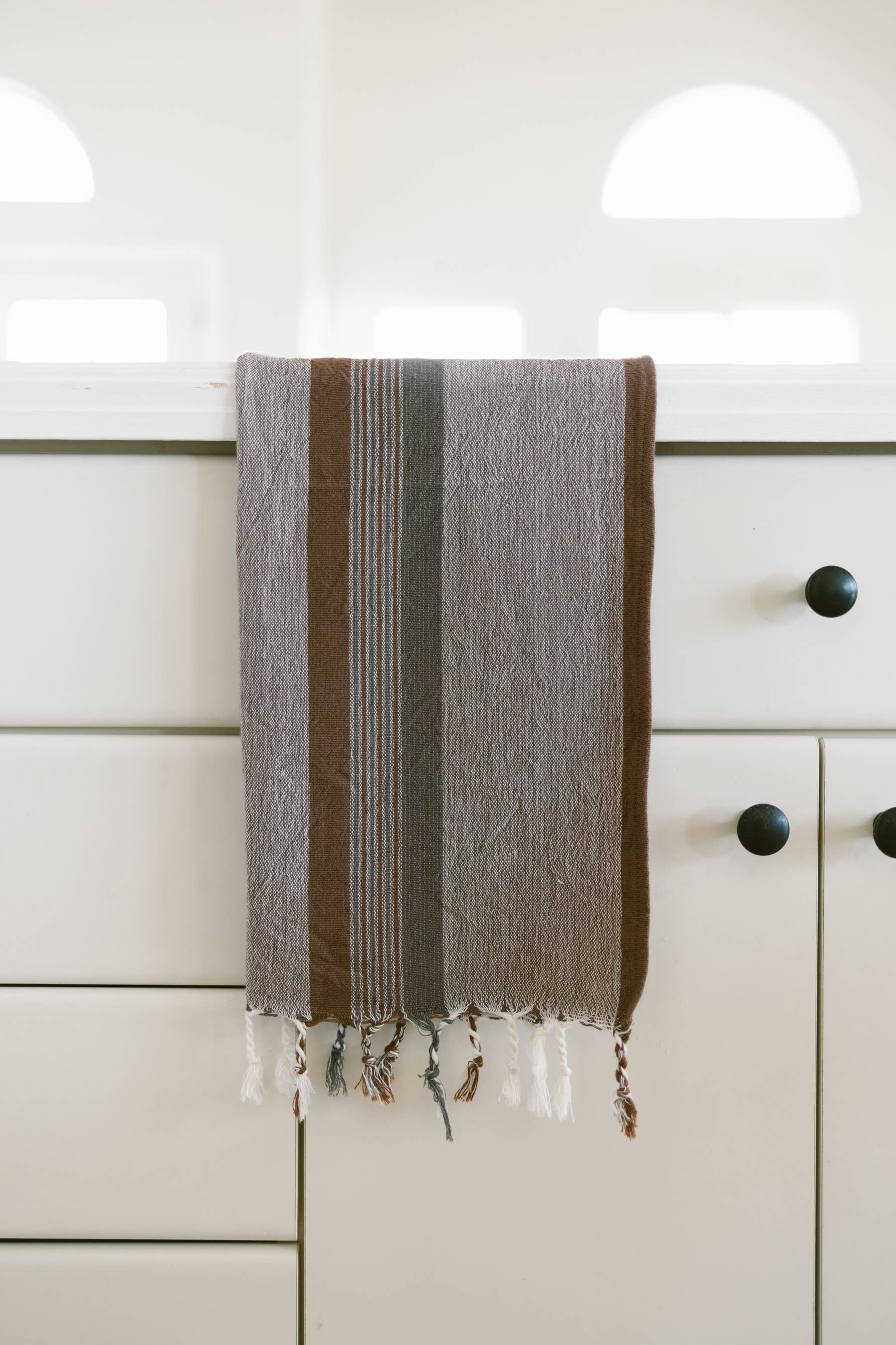 Grey, white, and dark brown cotton kitchen towel with fringe hangs on the door of a white kitchen cabinet with black knobs
