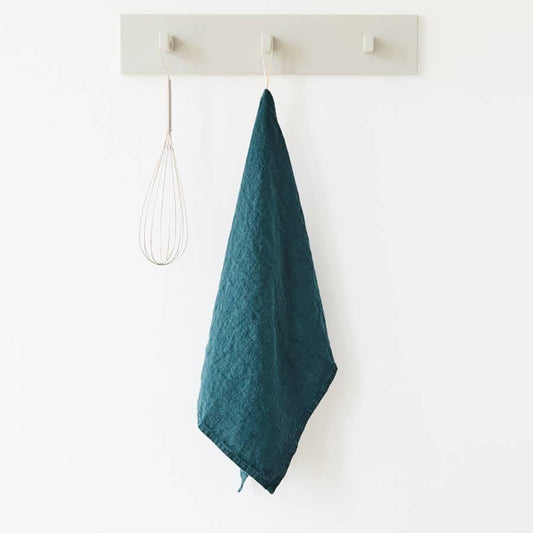 Teal linen kitchen towel hanging by it's loop on a hook next to a metal whisk against a white background