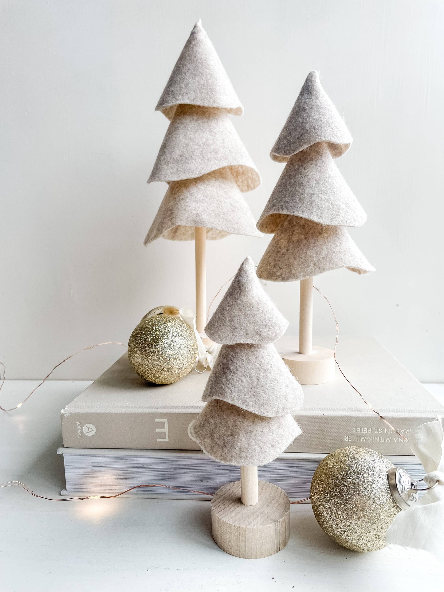 Three taupe felt Christmas trees on a washed wood base sit on a book with decorative holiday ornaments