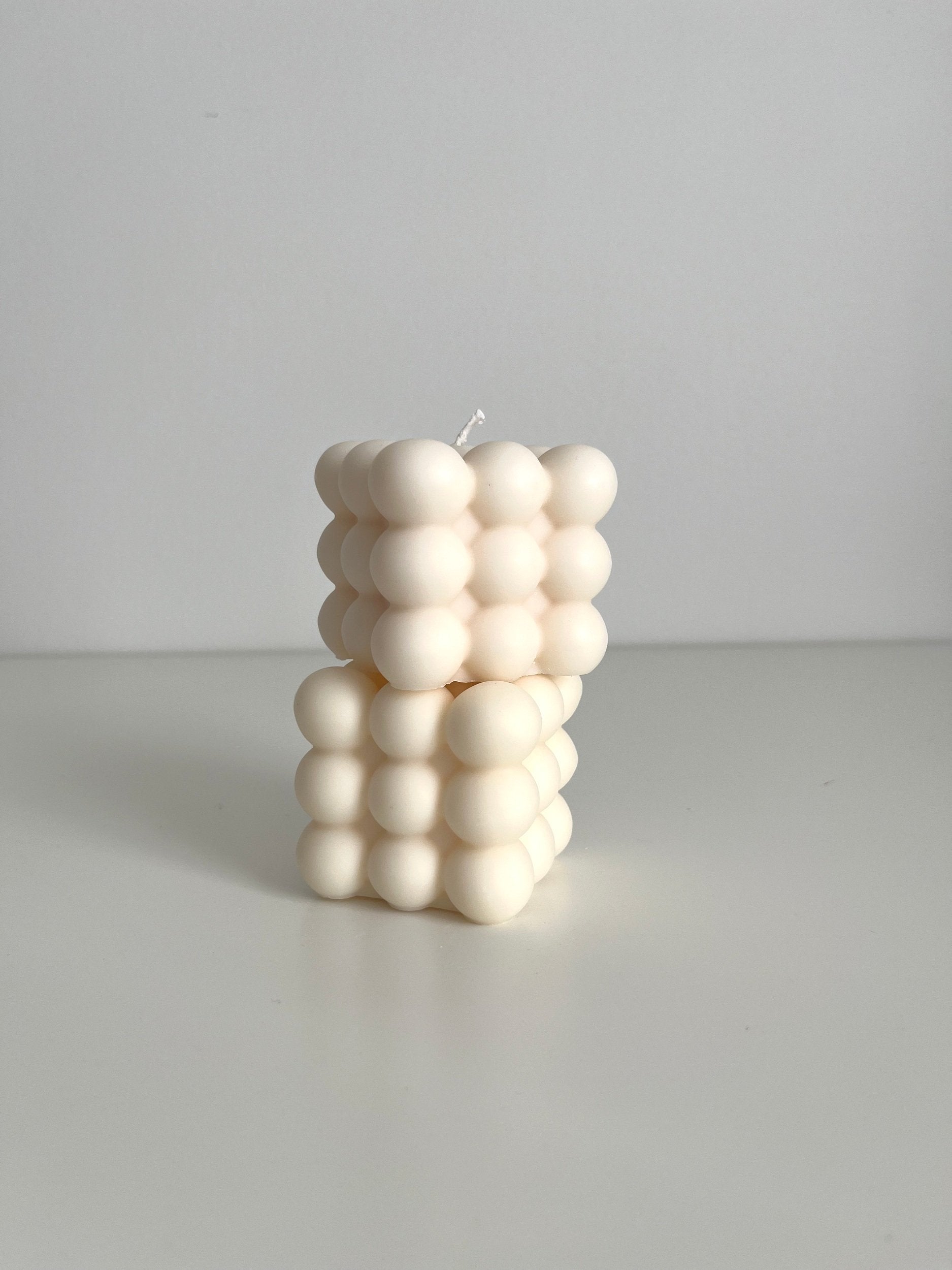 Two ivory bubble cube candles shown sitting stacked one above the other on a  white surface with a soft gray background