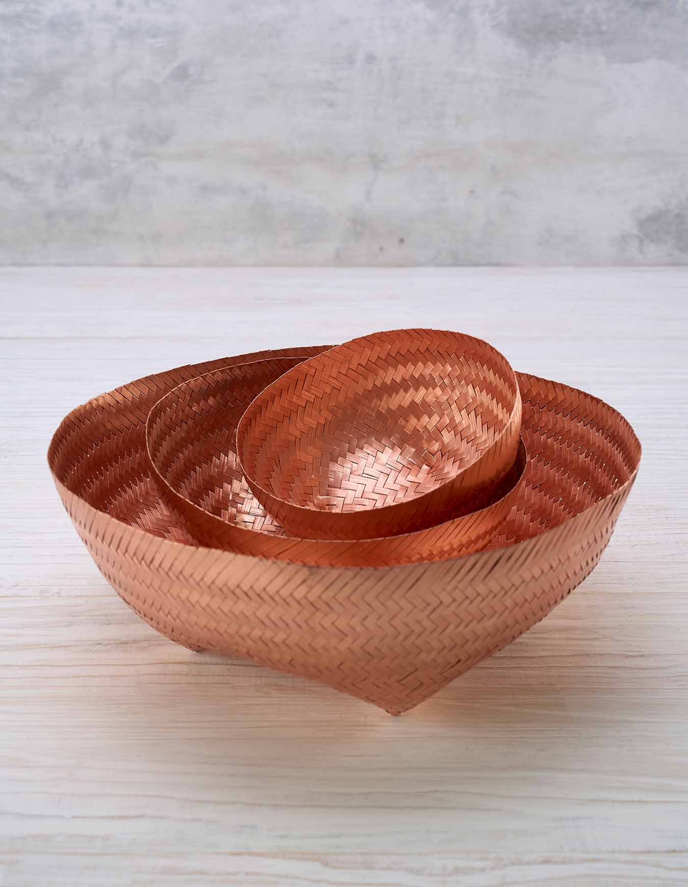 Three round woven copper baskets - small, medium, and large - sit nested within each other on a white surface against a light grey background