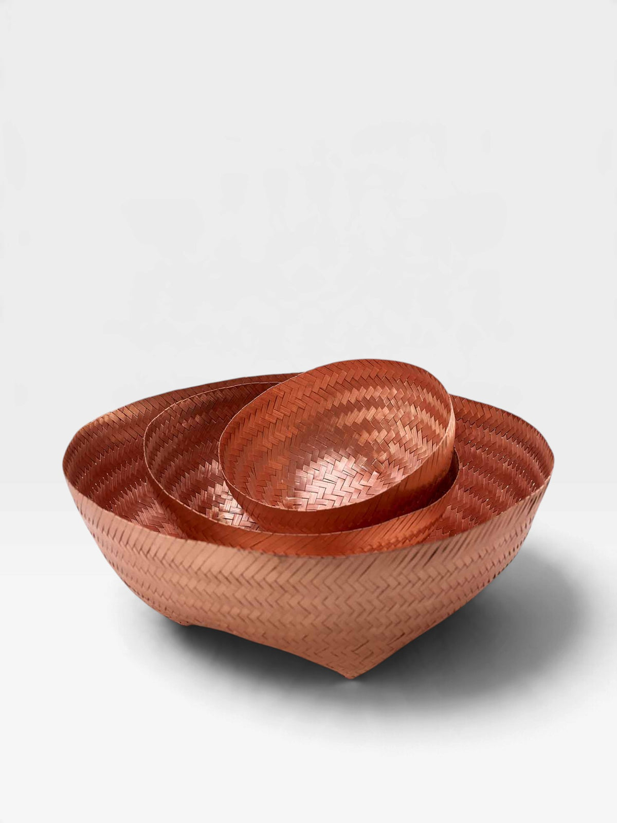 Three nested woven bowls in a warm copper tone, photographed on a clean light gray studio background.