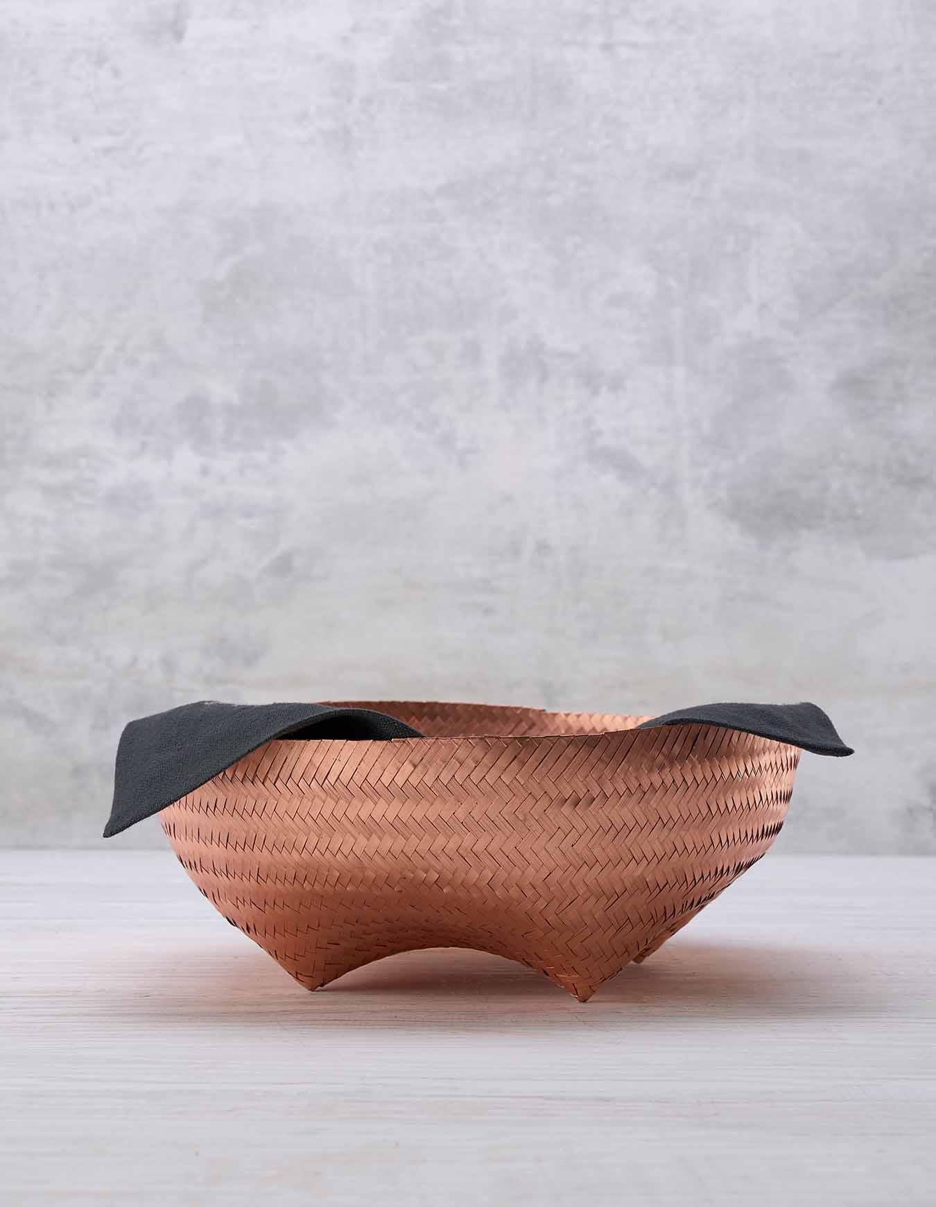 A footed copper woven basket with a black cloth napkin lining the interior sits on a white surface against a light grey background
