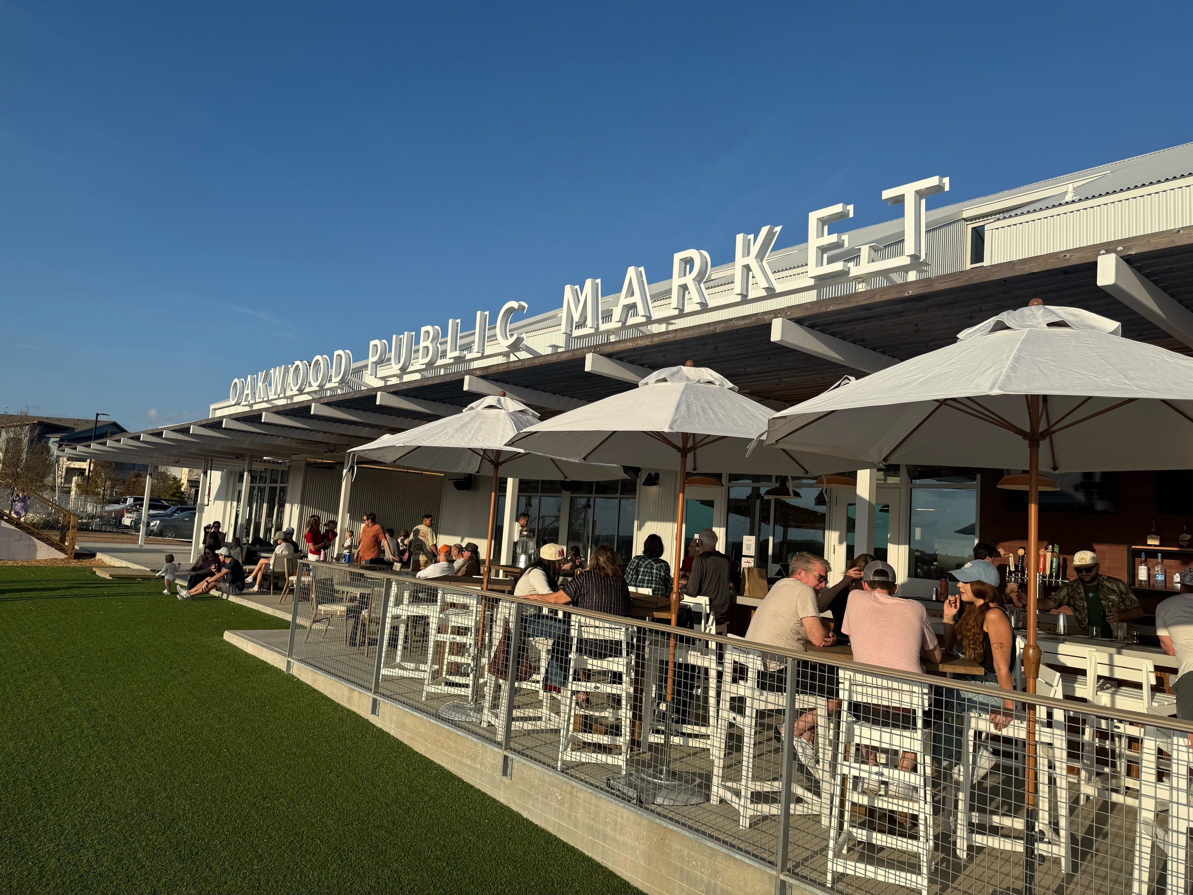 People sitting under white umbrellas at an outdoor seating area with 'Oakwood Public Market' sign.