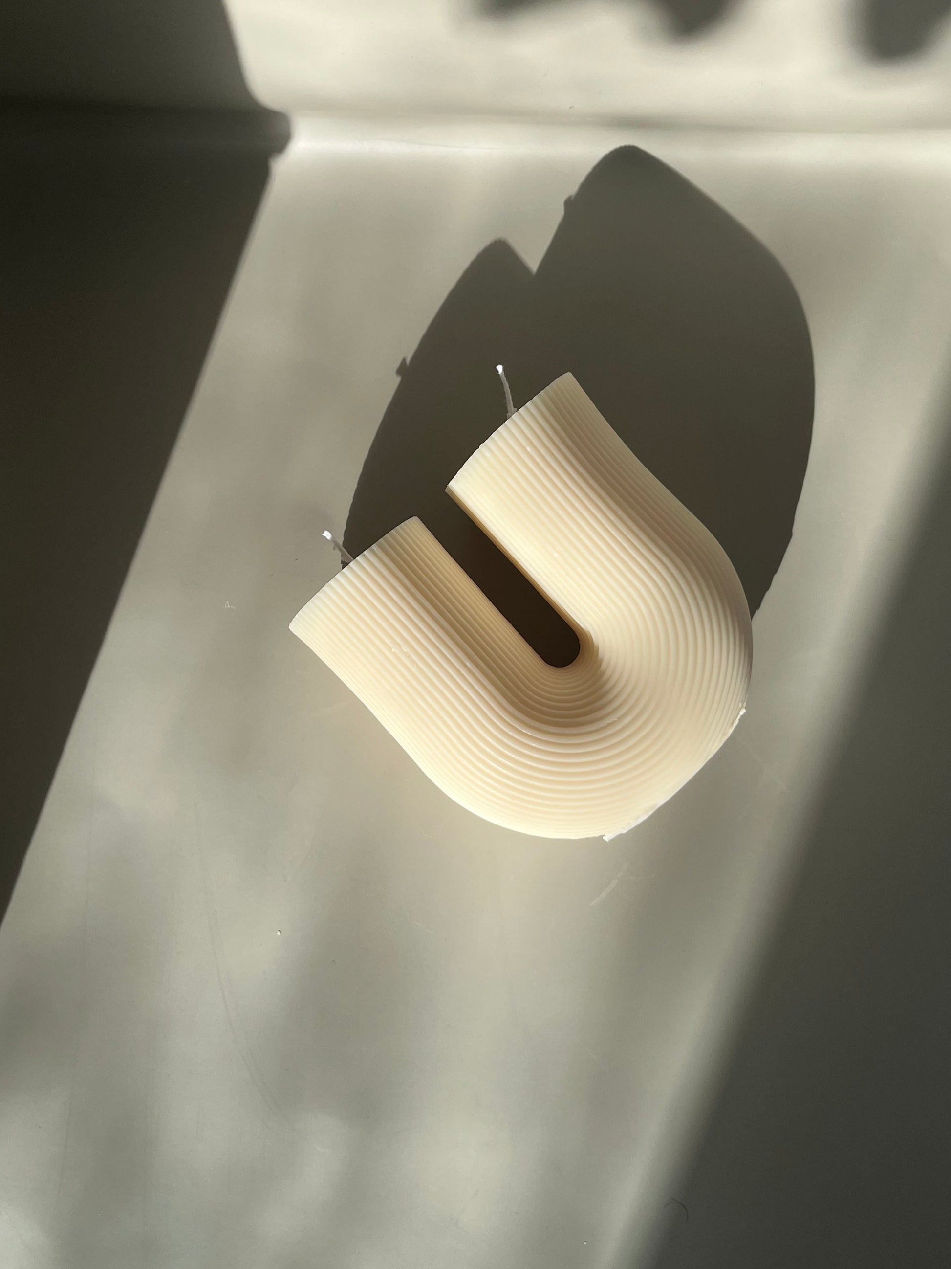 A u-shaped wax candle with ribbed detailing laying flat on an off-white surface with shadows