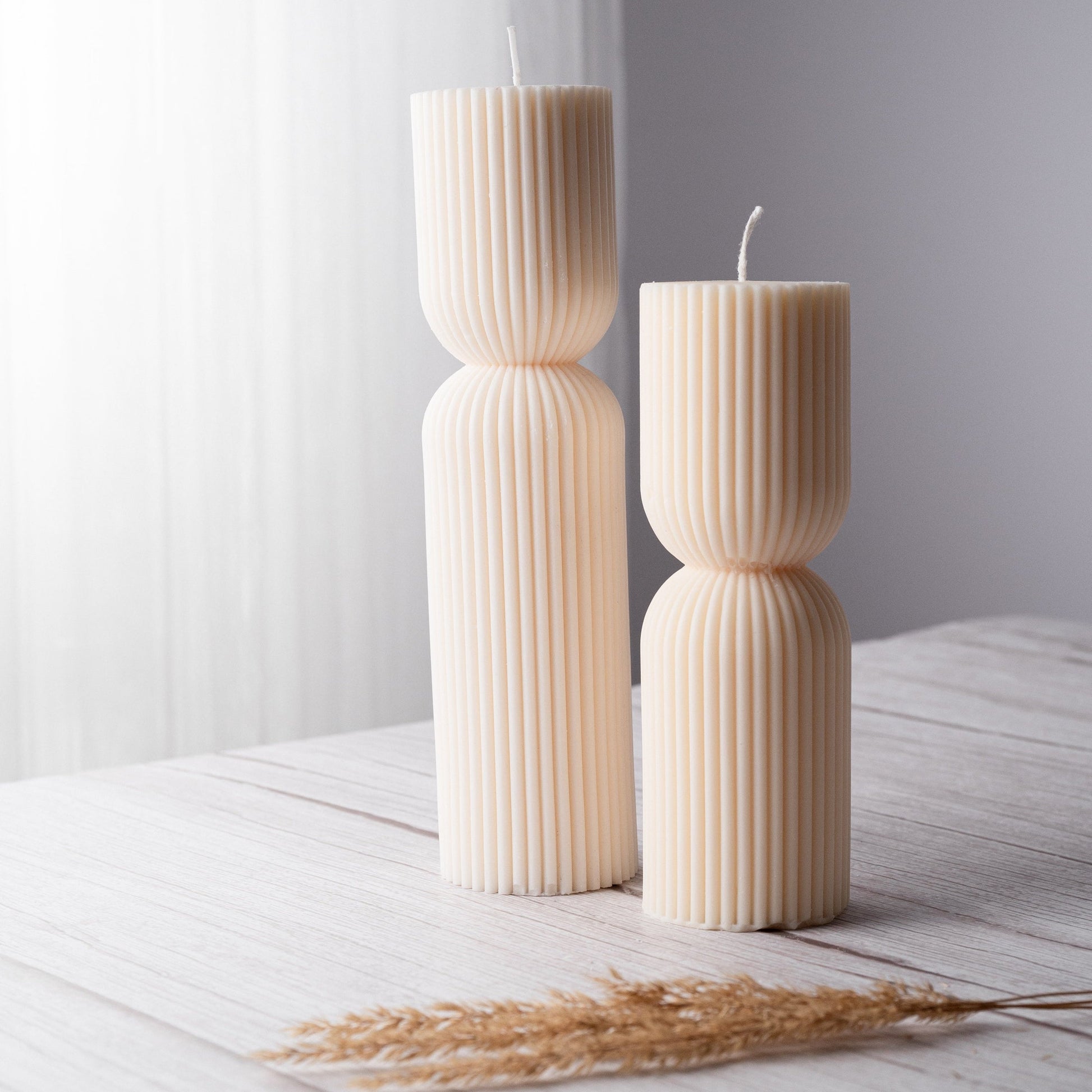 Two ivory pillar candles with ribbing and a cinched middle stand side by side on a white wooden surface with a dried brown plant stalk laying in front of them