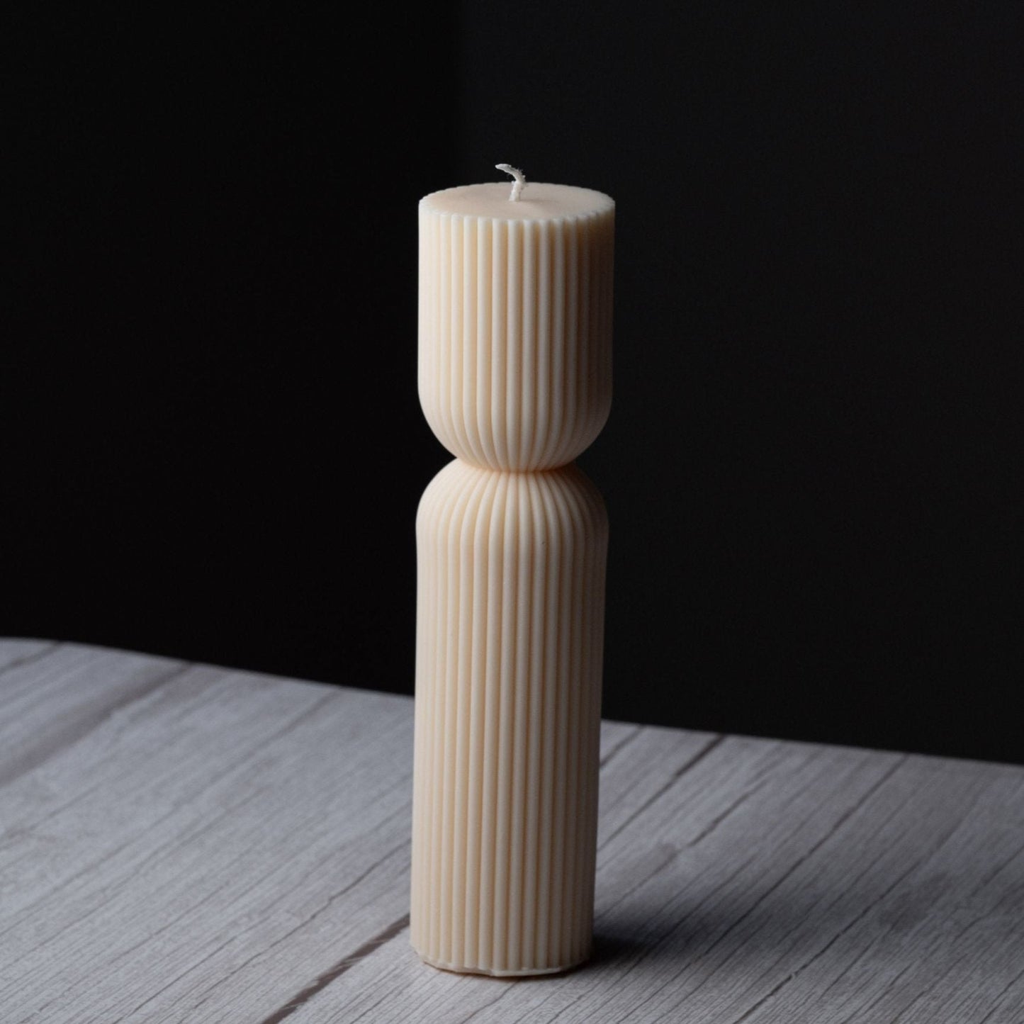 A tall ivory pillar candle with ribbing and a cinched middle stands on a gray wooden surface with a black background