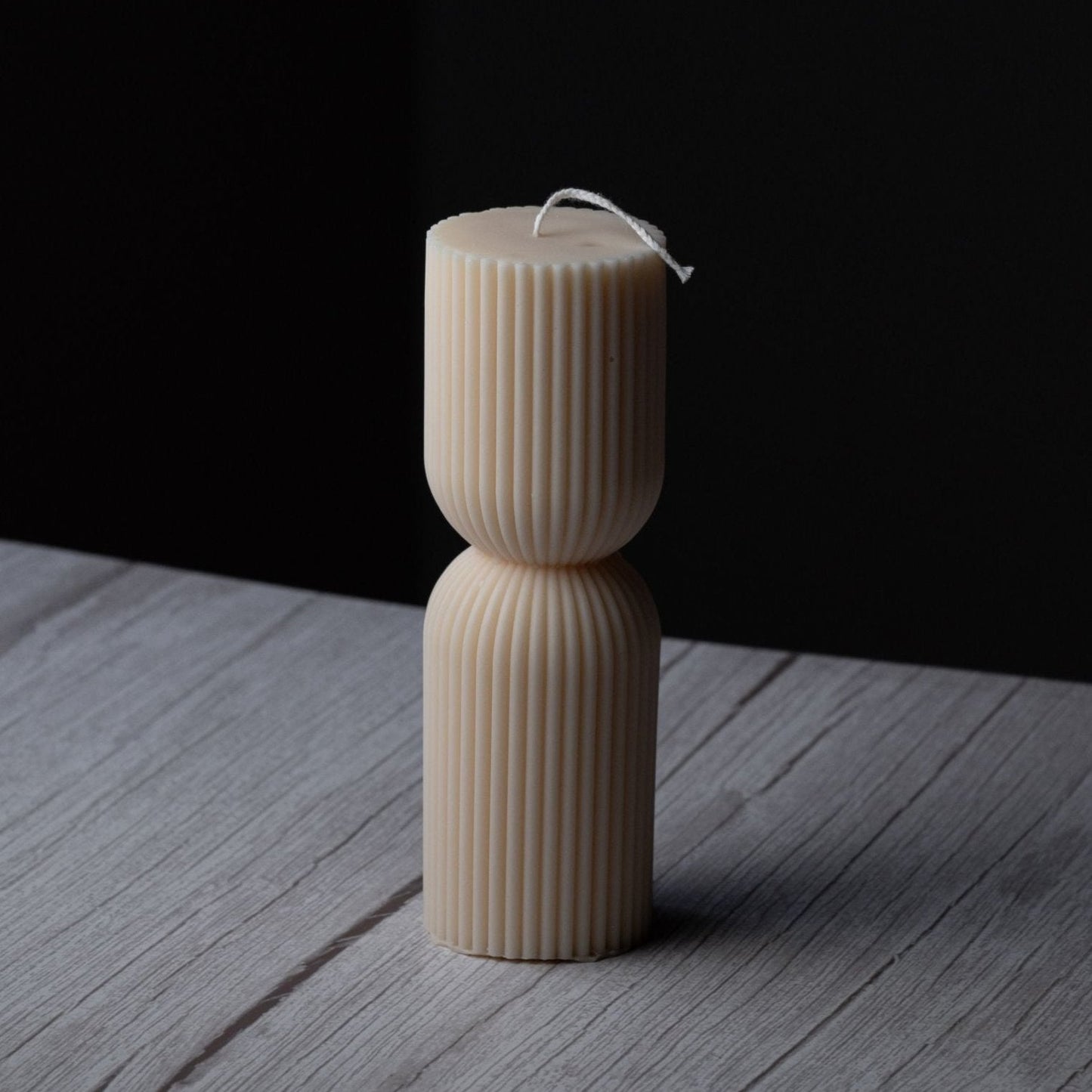 A small ivory pillar candle with ribbing and a cinched middle stands on a gray wooden surface with a black background