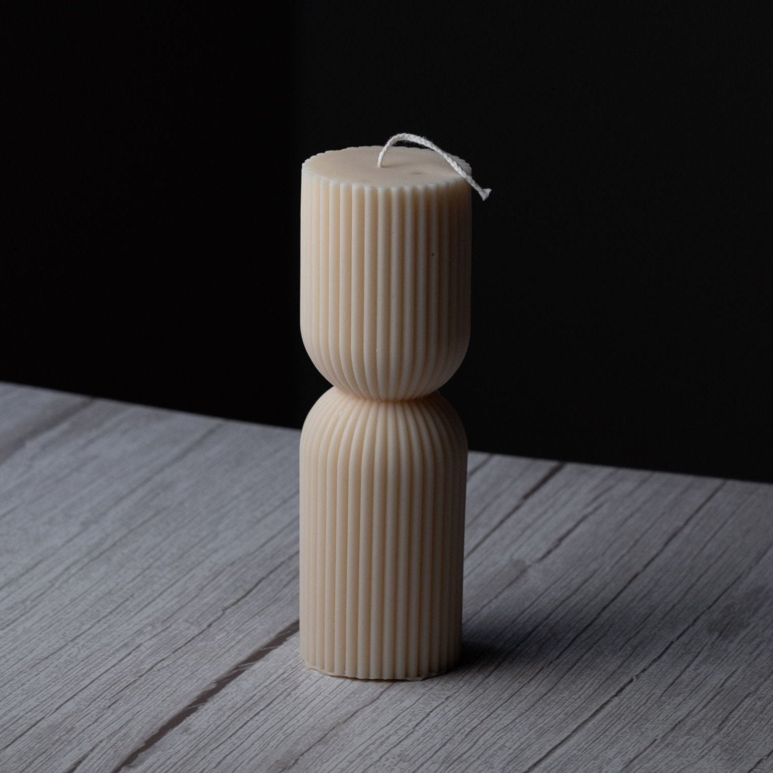 A small ivory pillar candle with ribbing and a cinched middle stands on a gray wooden surface with a black background