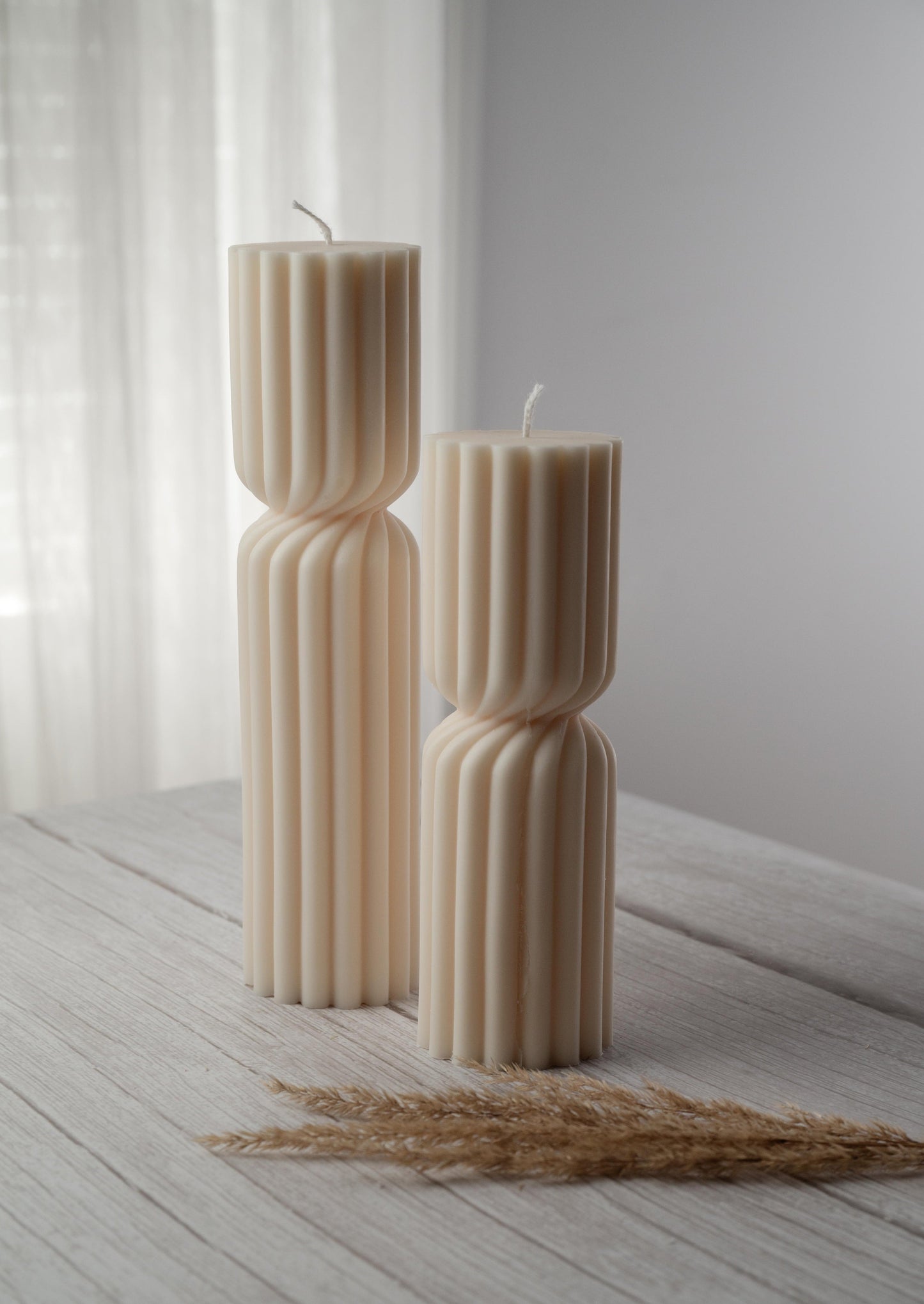 Two ivory candles of varying heights, a ribbed design, and a cinched twist at their middle sit side by side on a whitewashed wood surface