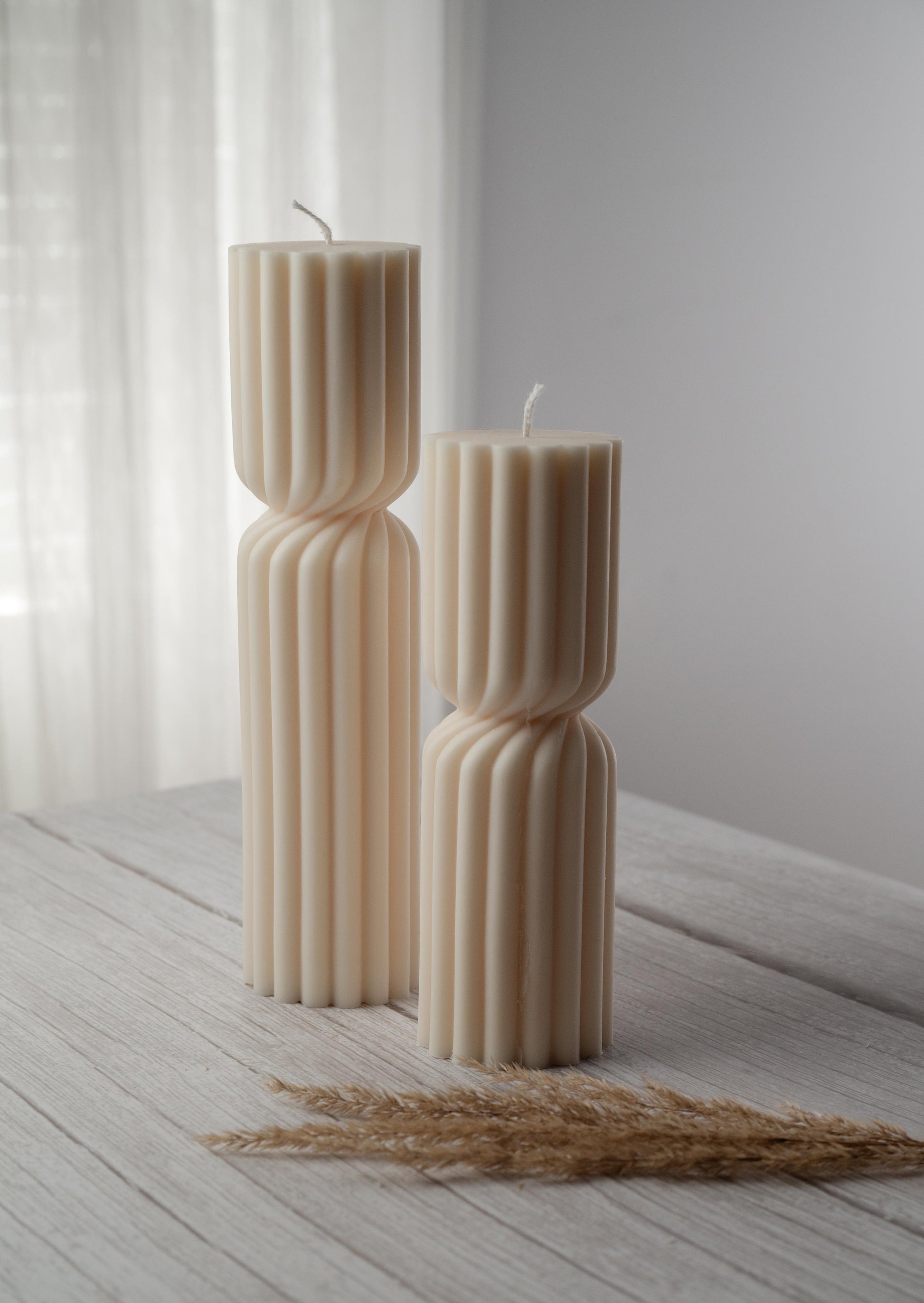 Two ivory candles of varying heights, a ribbed design, and a cinched twist at their middle sit side by side on a whitewashed wood surface
