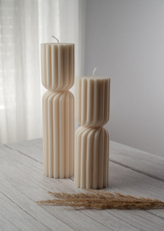 Two ivory candles of varying heights, a ribbed design, and a cinched twist at their middle sit side by side on a whitewashed wood surface
