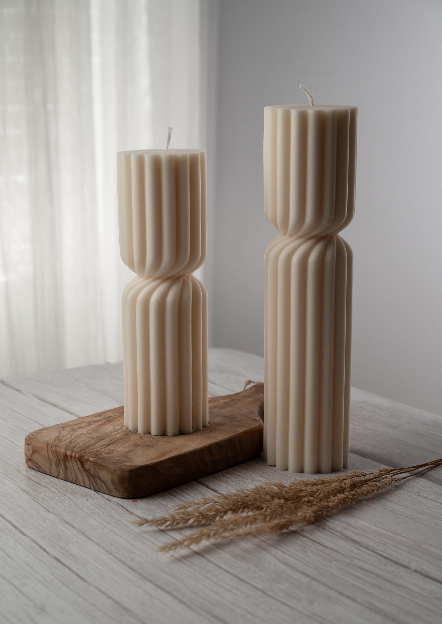 Two ivory candles of varying heights, a ribbed design, and a cinched twist at their middle sit side by side on a whitewashed wood surface, with the candle in the background resting on a paddle-shaped wooden board 