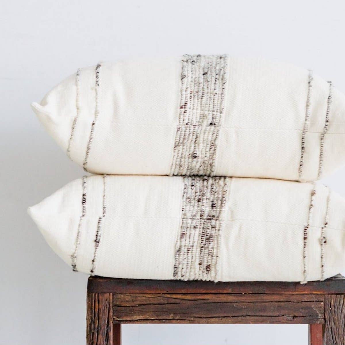 Two pillows laid on on top of one another, sitting on a wooden stool - both pillows have ivory wool and cotton covers with striping detail, one in ivory, the other in dark grey