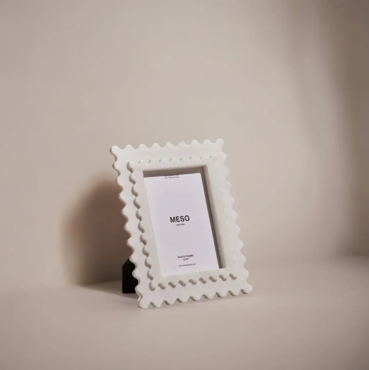 A white marble picture frame with scalloped edging set against a beige backdrop