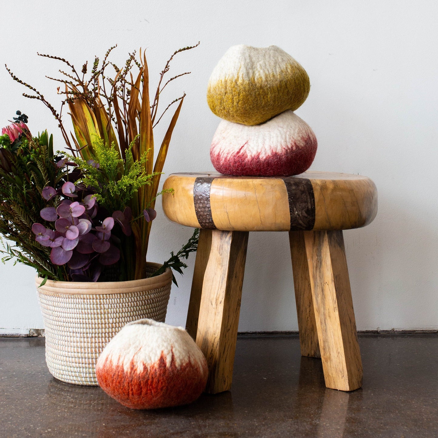 A wooden stool is shown with a potted plant beside it, two felted wool bowls sitting on top of it, and a third felted wool bowl sitting in front of it, by it's left leg