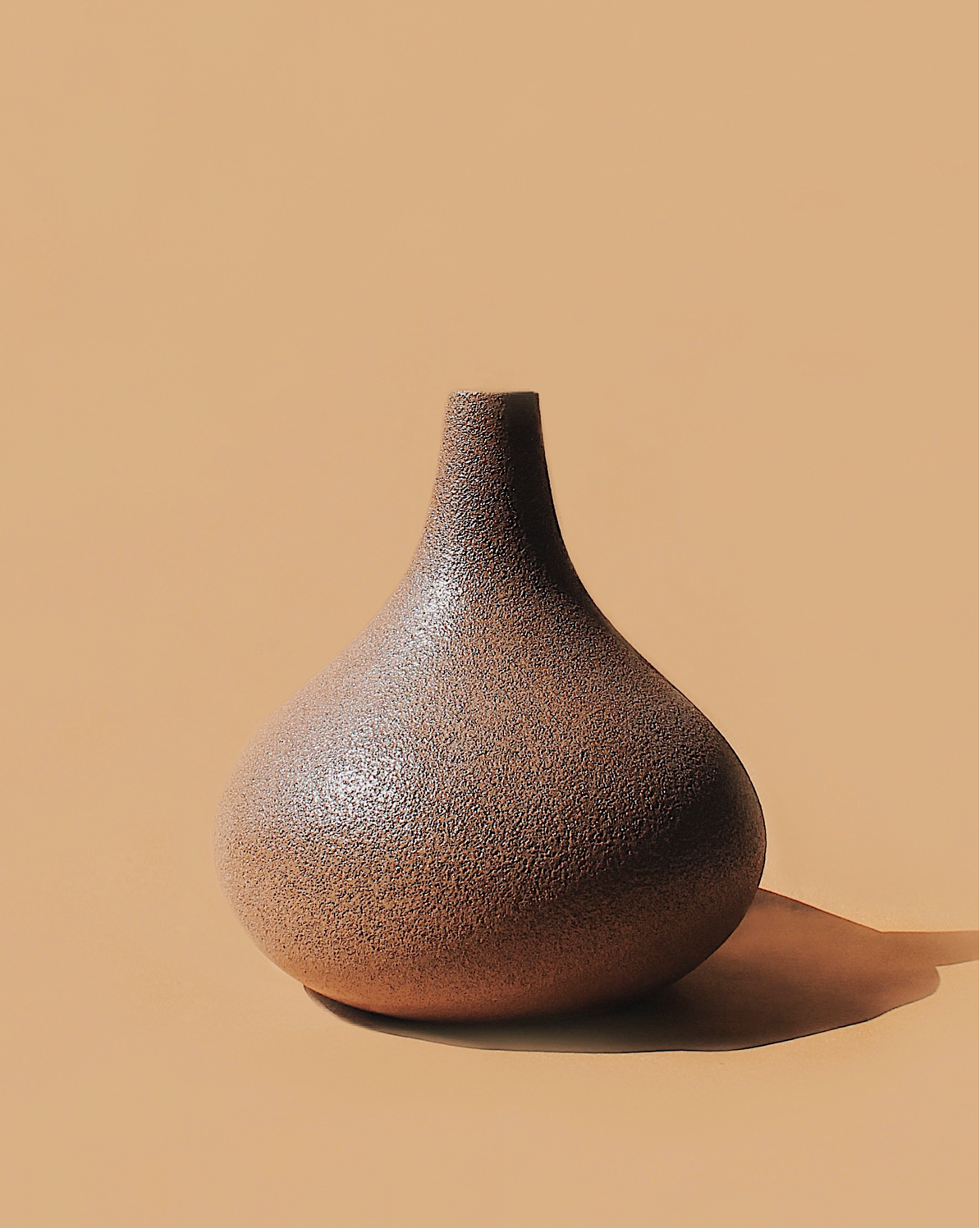 A garlic-shaped vase with terracotta and fine black speckles finish sits against a beige background